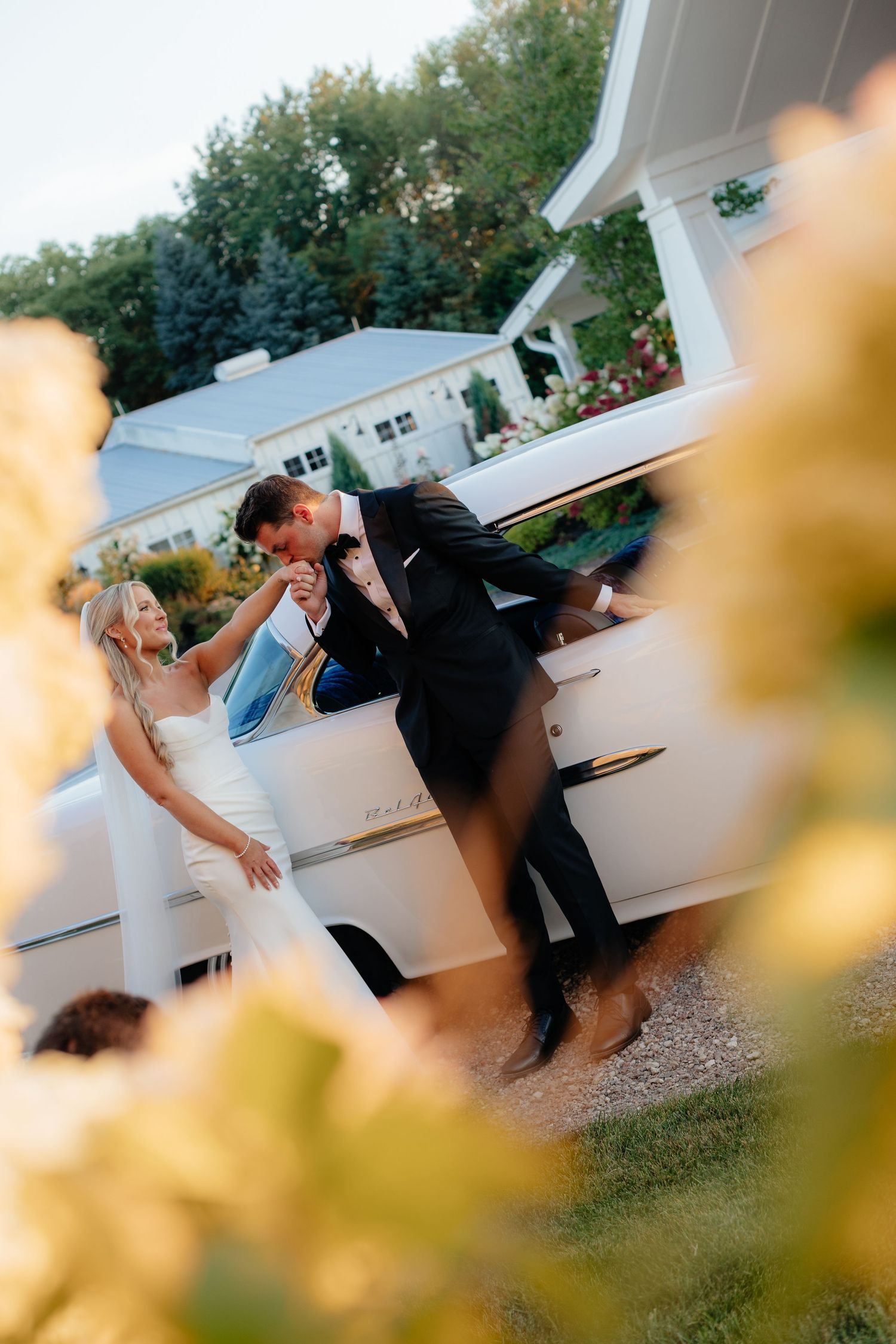 Elegant wedding couple poses playfully by a classic white car during golden hour at their outdoor celebration.