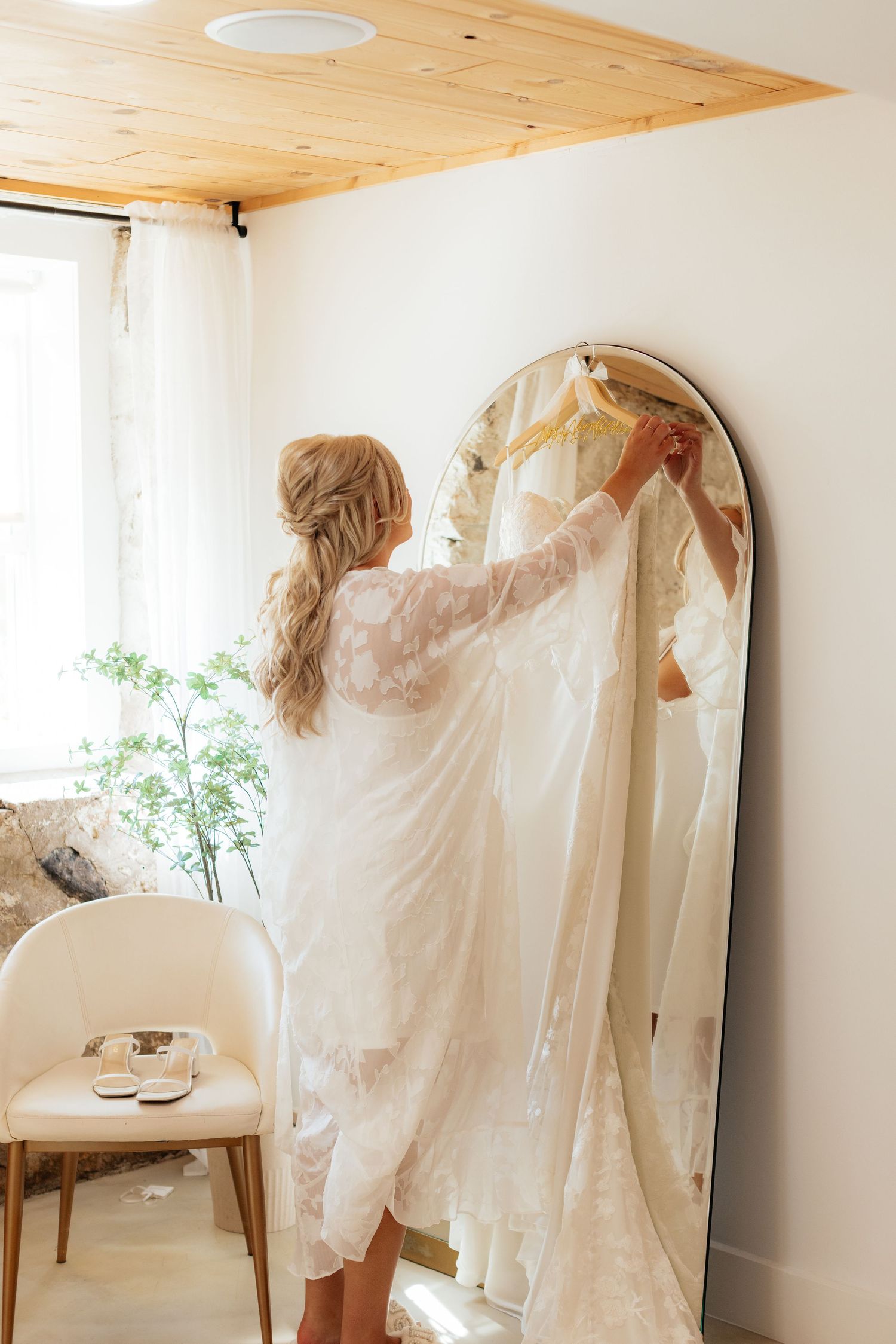 A bright dressing room shows a wedding dress being prepared near an arched mirror.