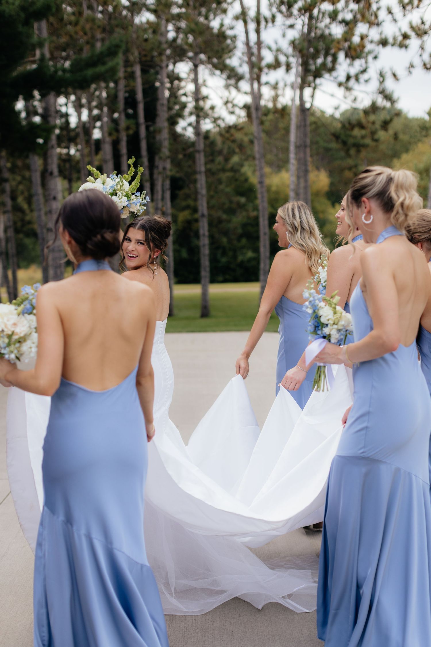 Bridesmaids in flowing periwinkle dresses help with the bride's train in an outdoor garden setting.