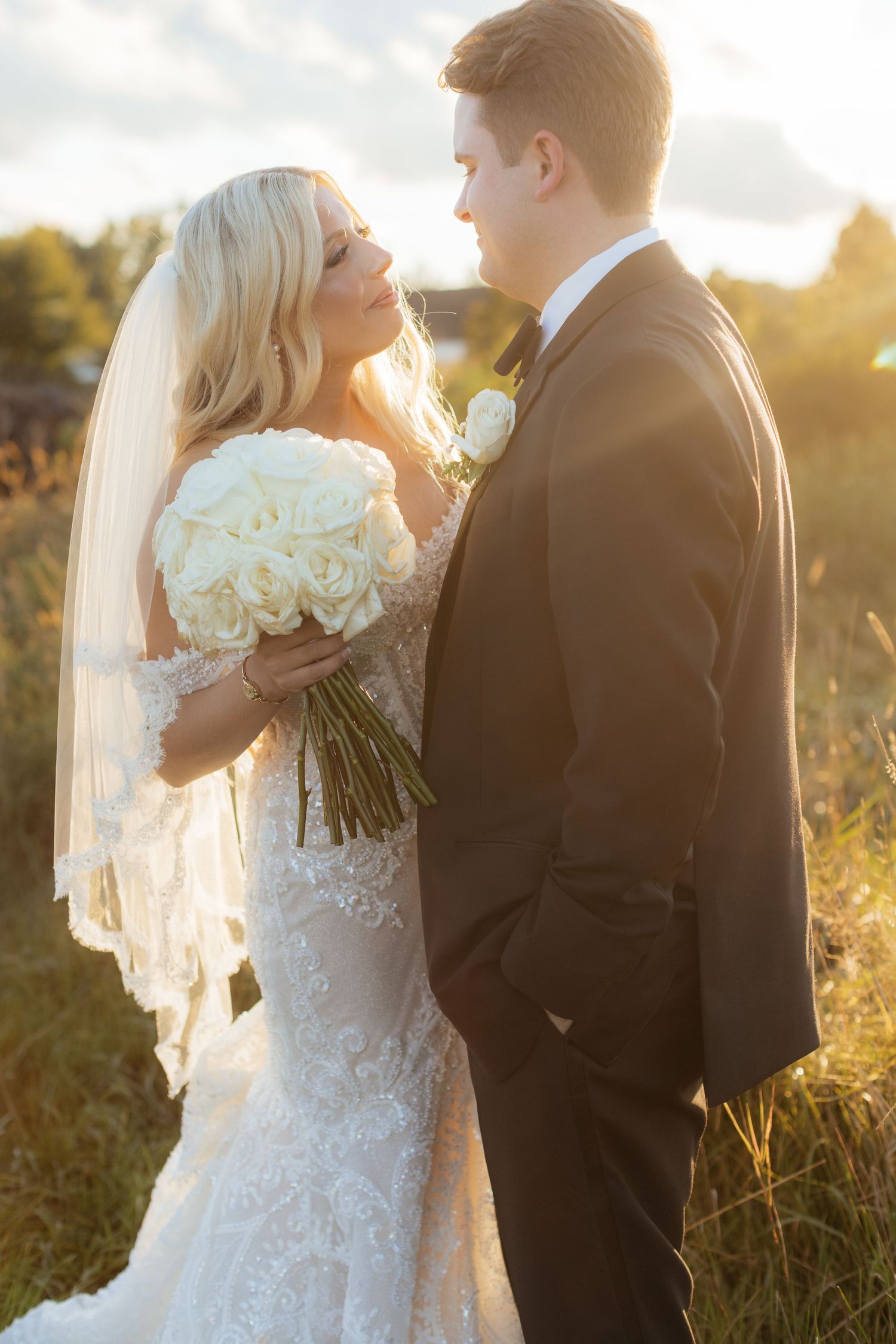 Couple in formal wedding attire embracing during golden hour in a field.