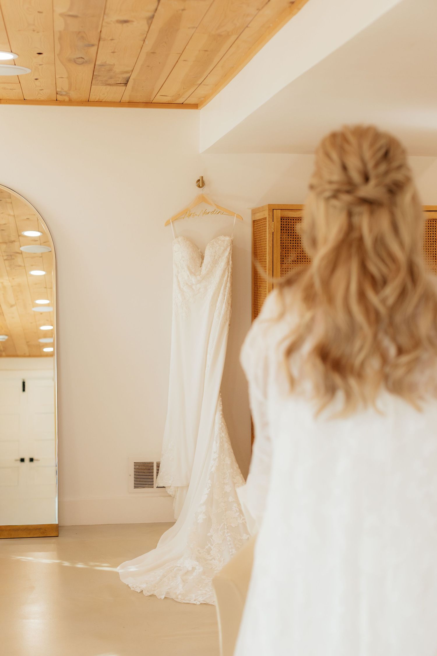 A wedding dress hangs in a bright room with wooden ceiling and white walls.