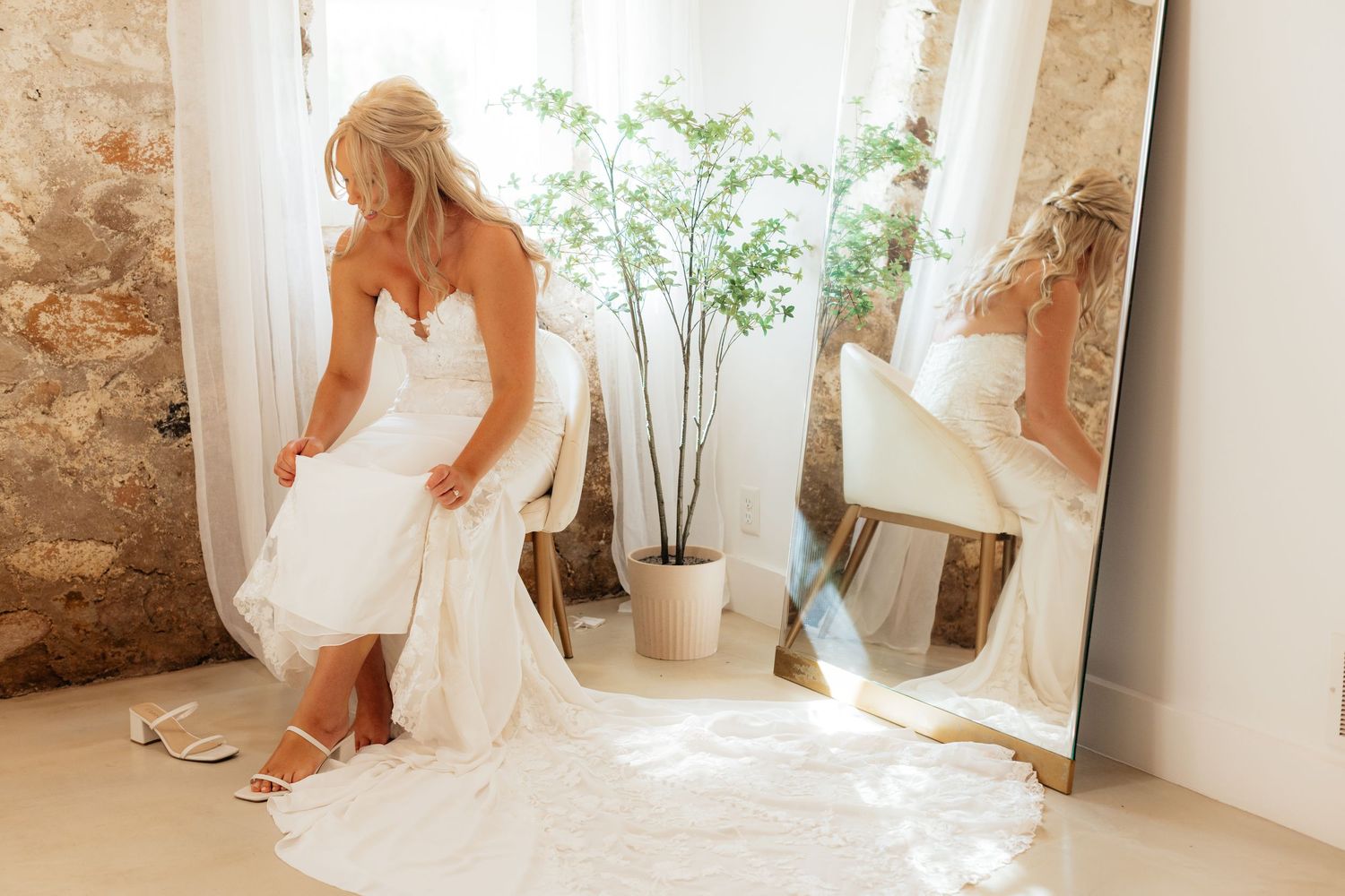 Elegant white wedding dress photographed in bright room with stone wall and natural lighting.