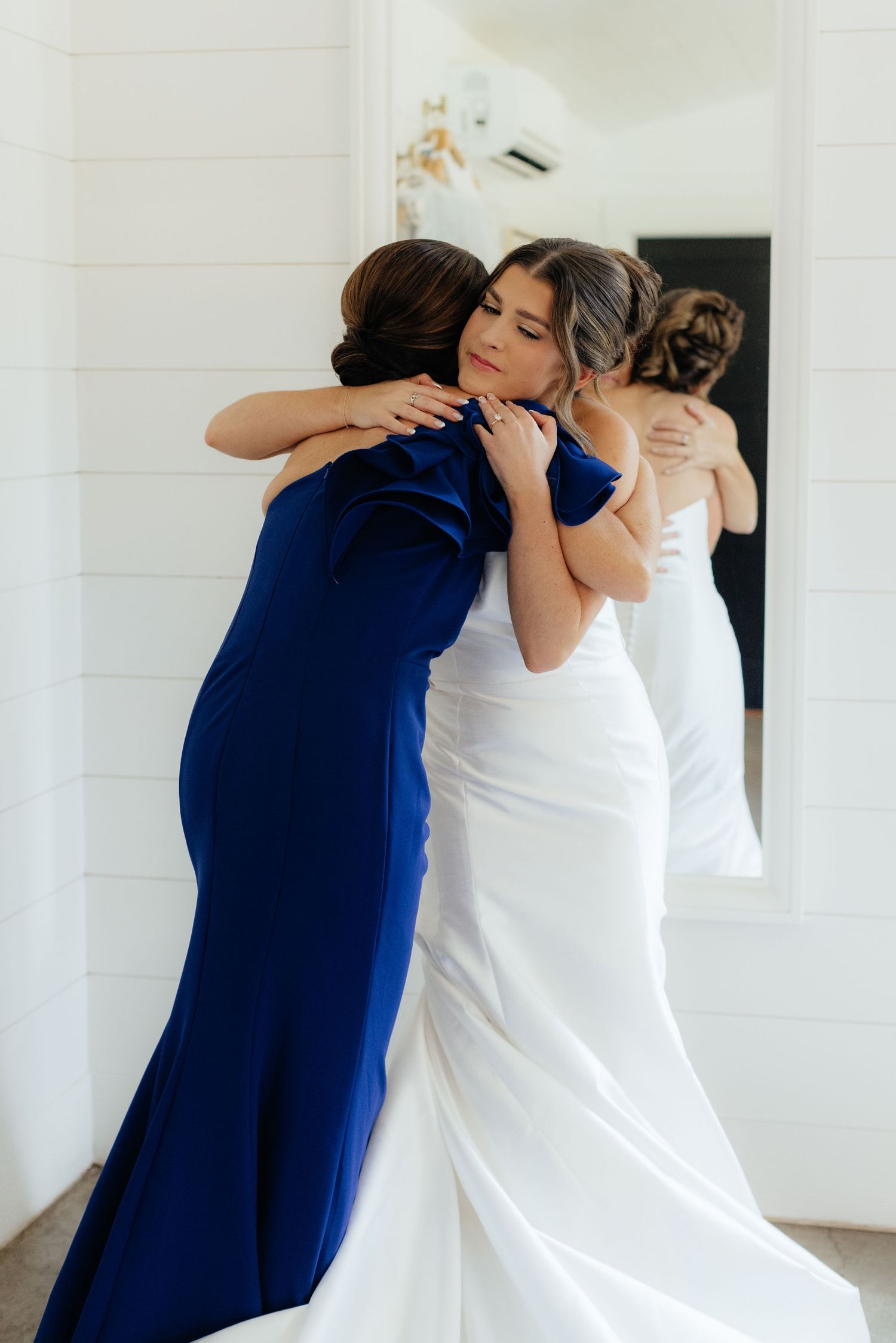 Emotional embrace between two women in white and navy dresses during wedding preparations.