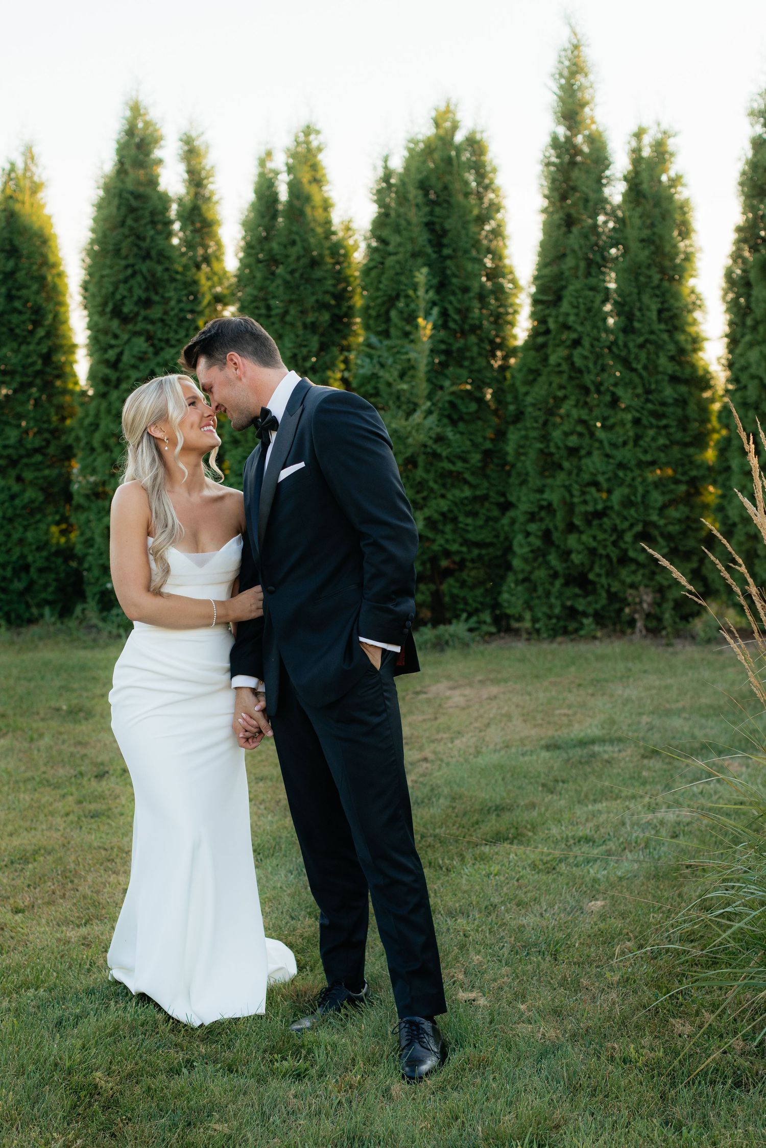 Bride and groom share intimate dance moves on lawn with row of cypress trees behind them.