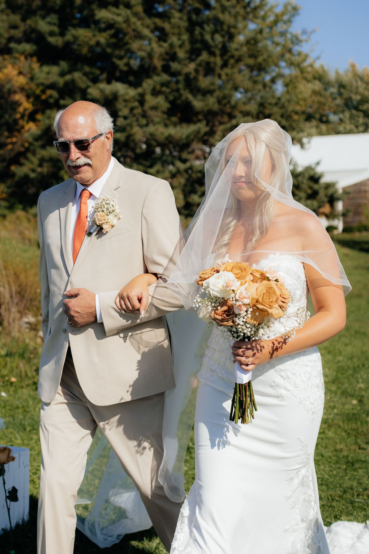 Wedding party members in beige suits and white dresses hold bouquets outdoors.