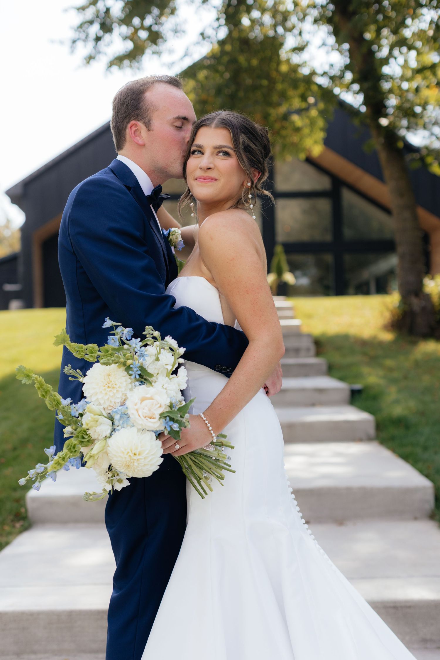 Newlyweds share intimate moments outdoors with white floral bouquet against rustic buildings.