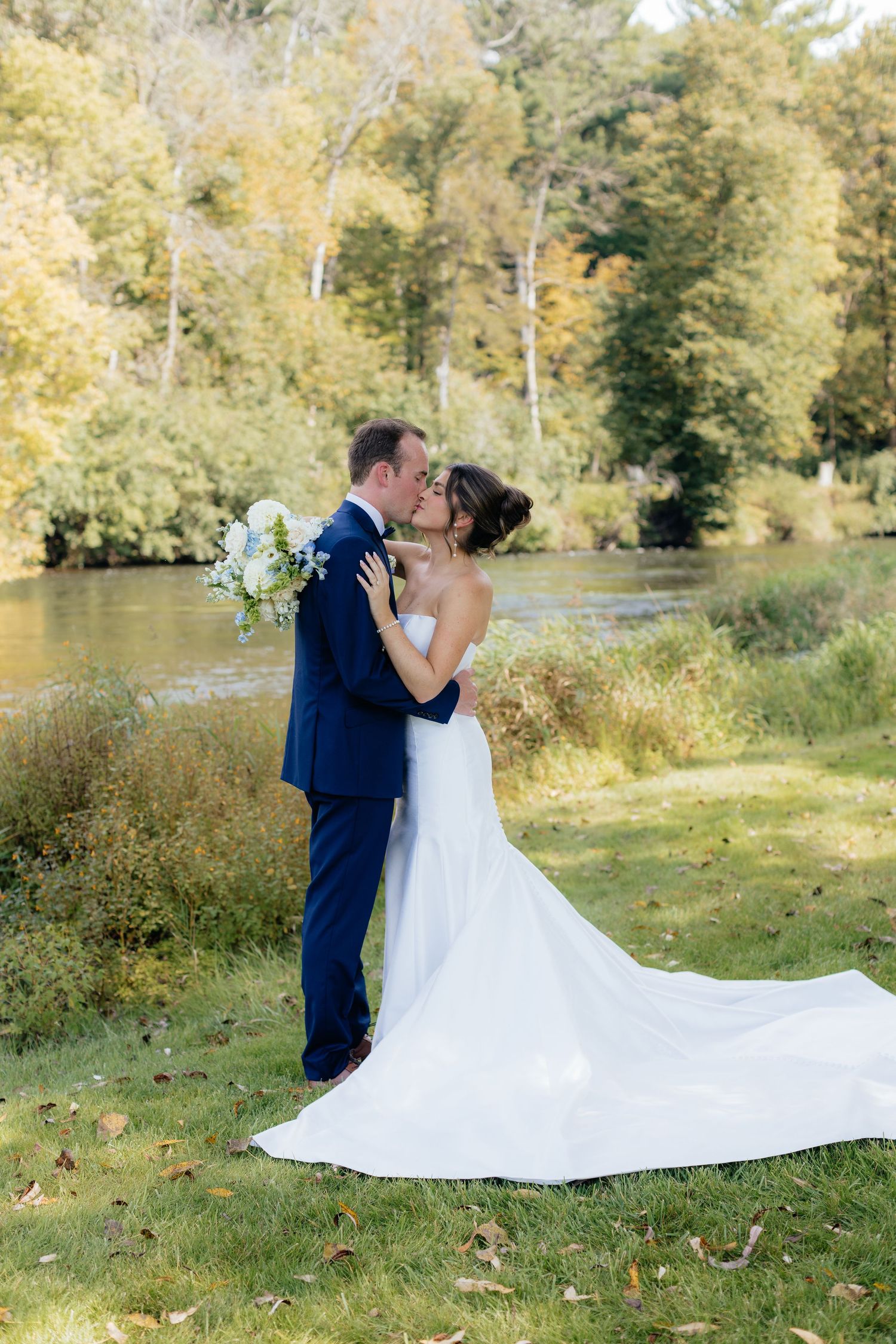 Bride in flowing white gown kisses groom by riverside during fall wedding.