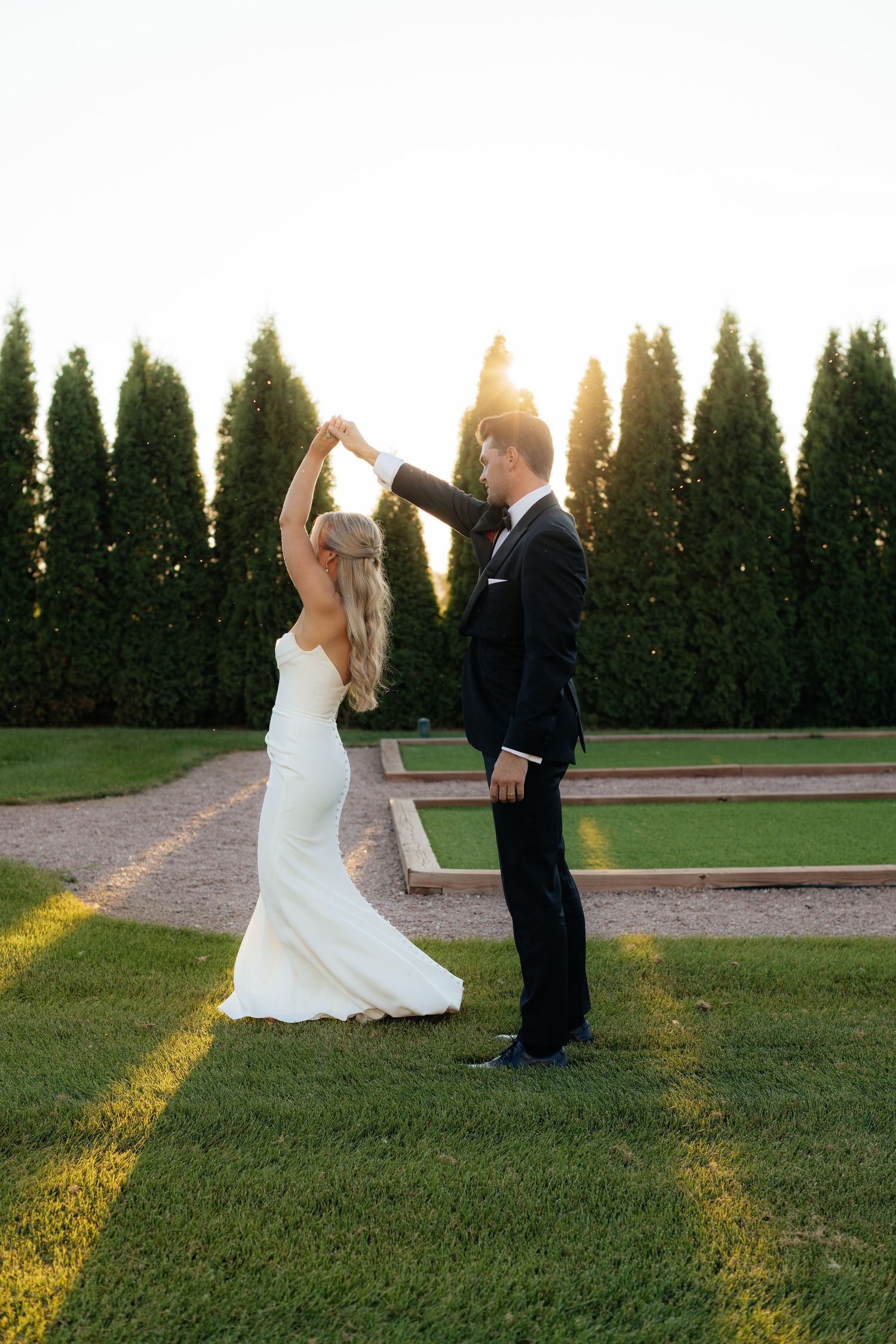 Newlyweds dance together on a grassy lawn with evergreen trees in the background at sunset.