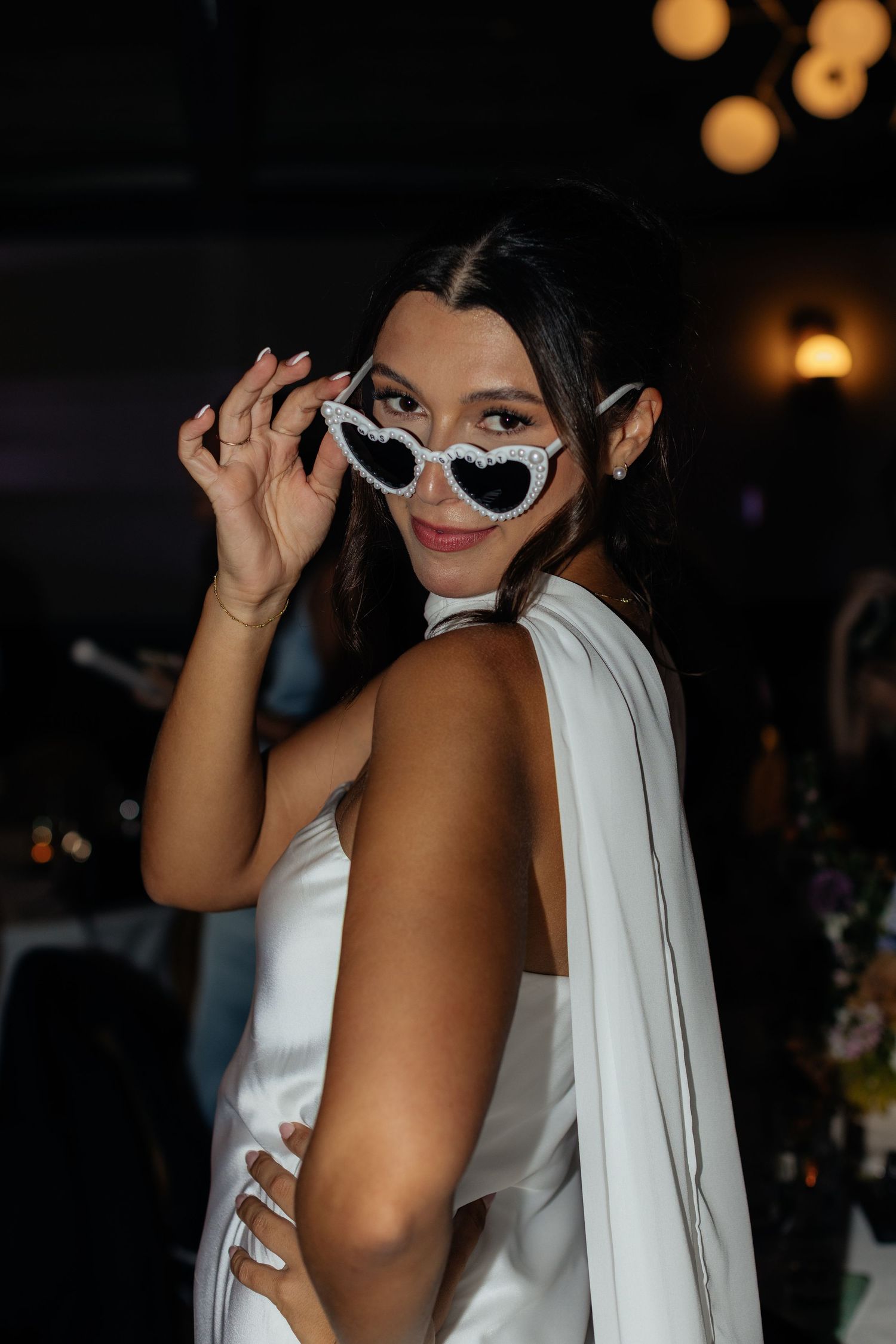 A person in a white dress poses playfully with heart-shaped sunglasses at a celebration.