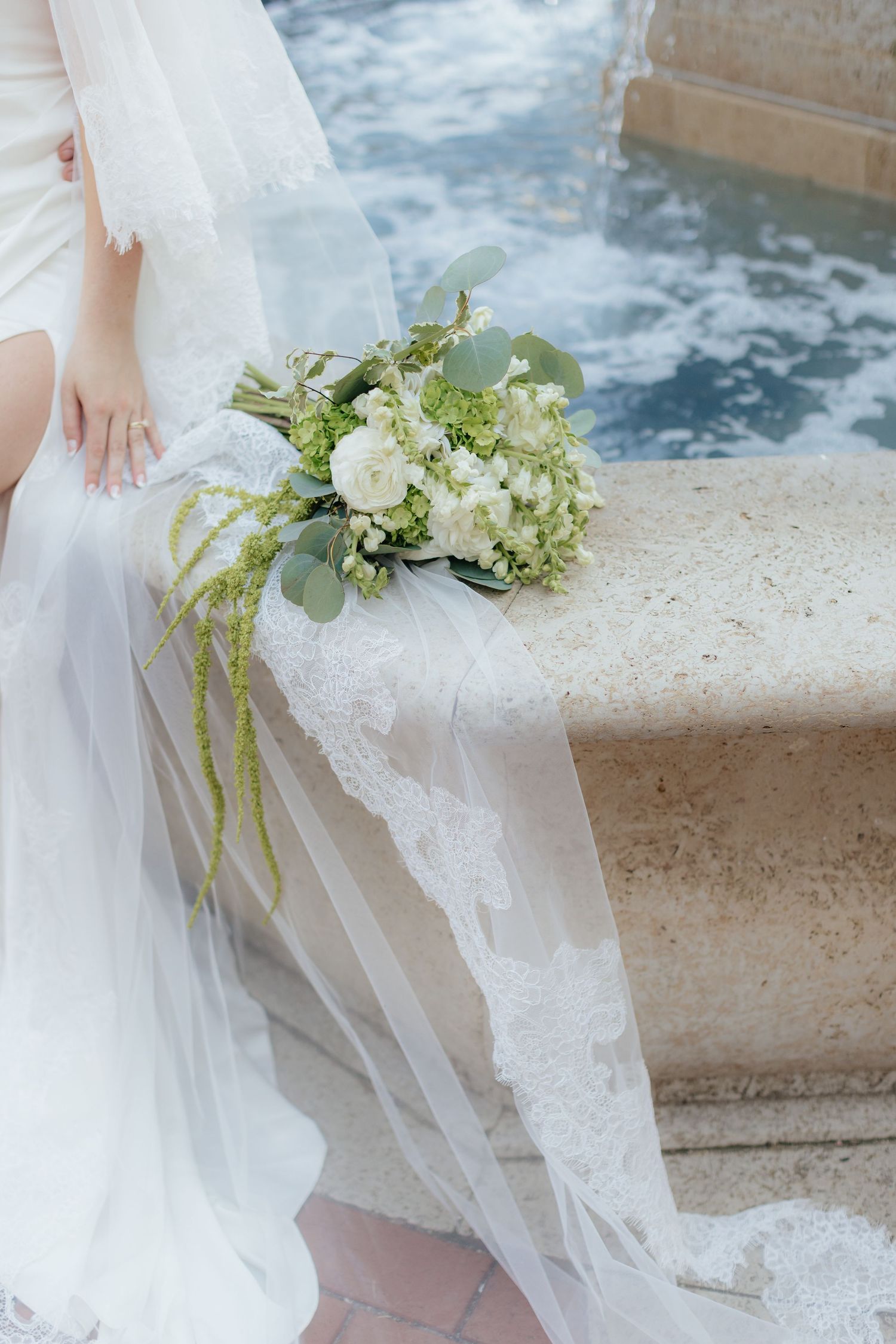 White and green bridal bouquet resting on stone fountain edge with flowing veil.