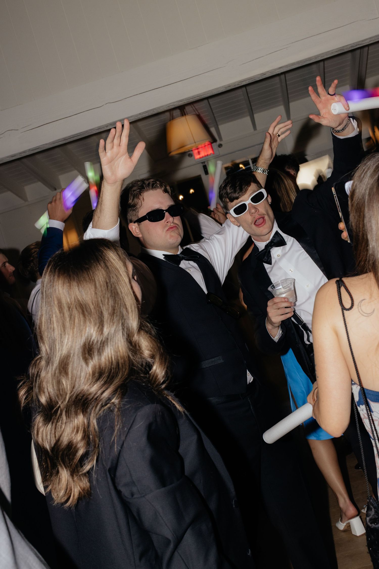 Wedding party guests dancing with sunglasses and glow sticks under dim lighting.