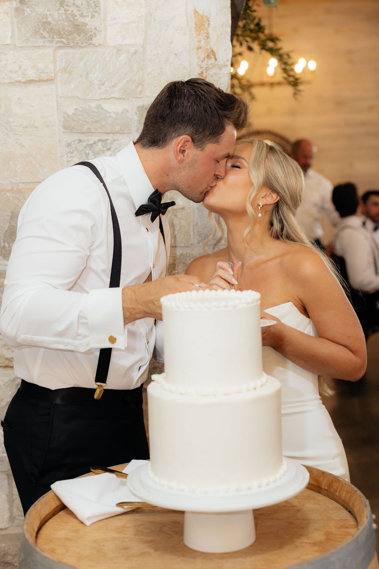 Newlyweds sharing a kiss while cutting their elegant two-tier white wedding cake.