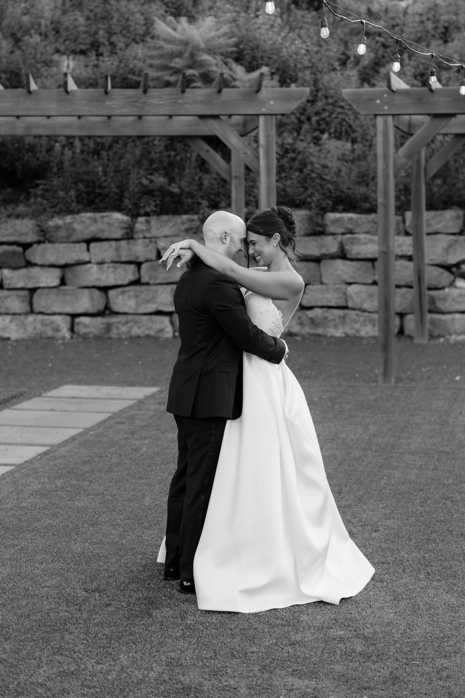 Black and white photo of newlyweds dancing on outdoor lawn near stone wall.