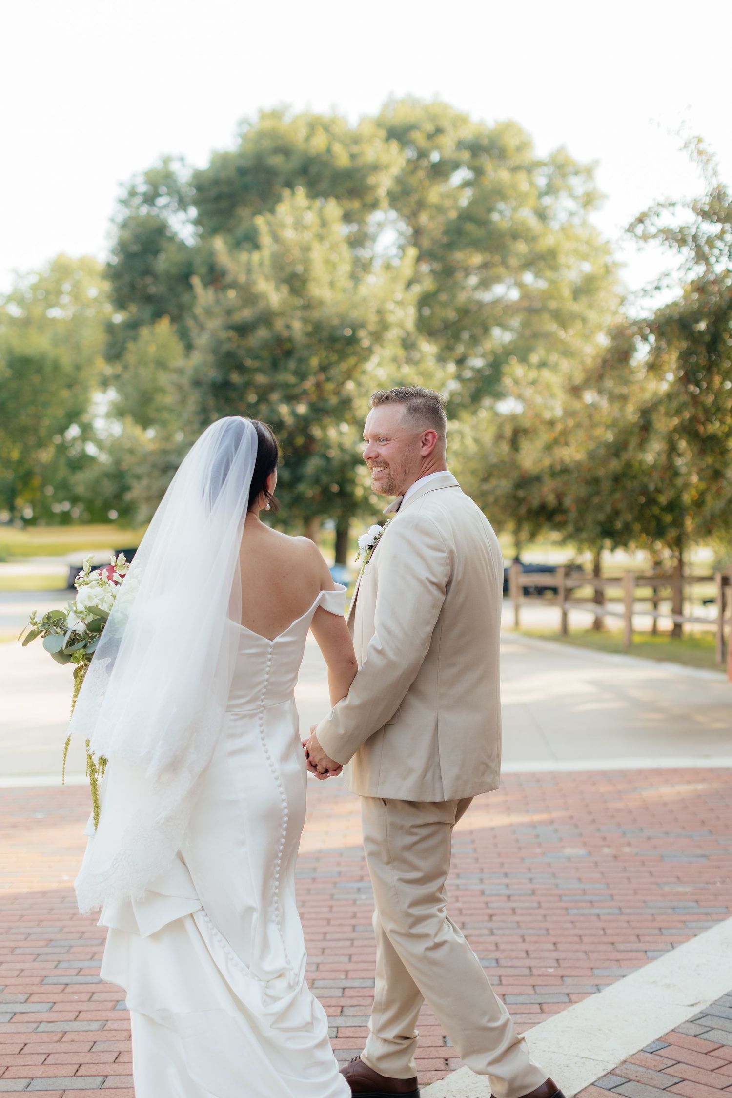 Bride and groom walking together on brick pathway surrounded by trees on their wedding day.
