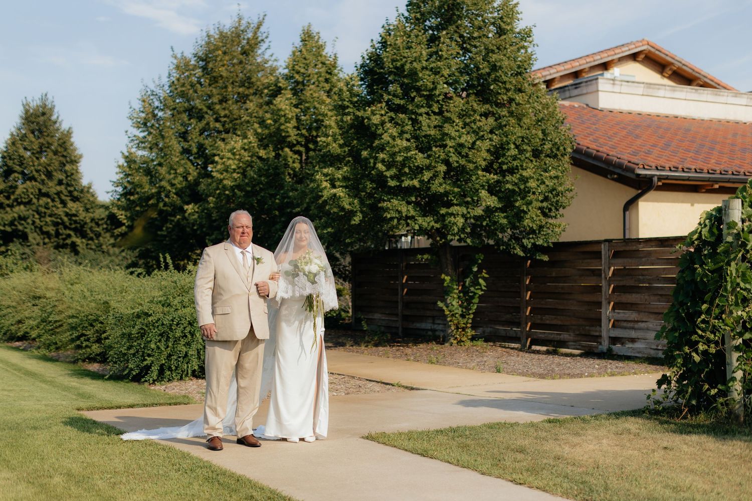 Wedding couple walks hand in hand across scenic outdoor venue with buildings in background.