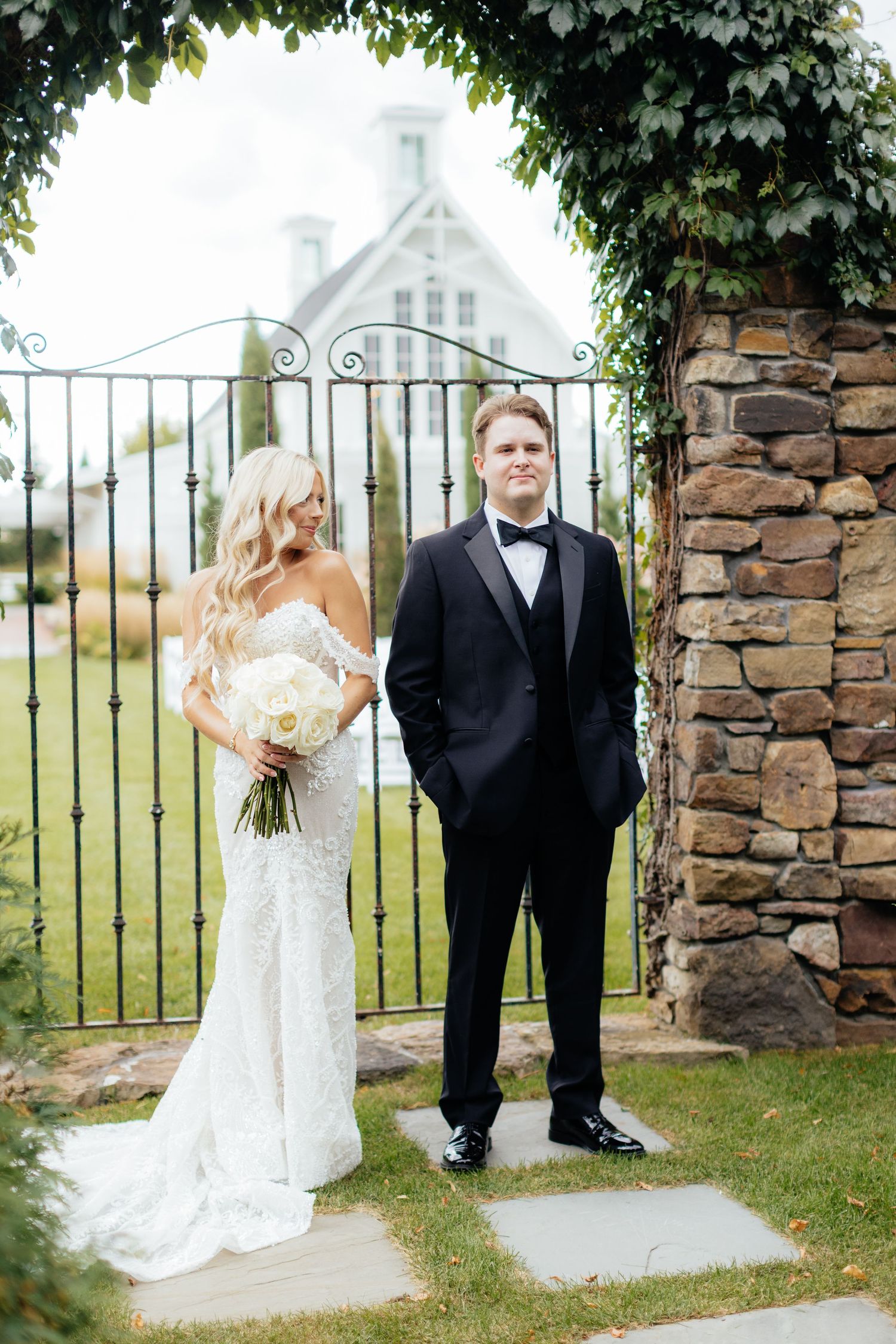 Newlyweds stand together by a rustic stone gate with a white chapel visible in the distance.