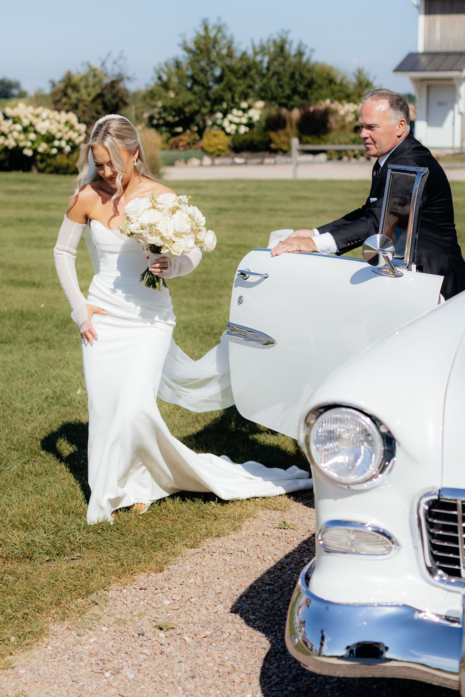 Elegant bride in white gown stands beside classic Chevrolet car.