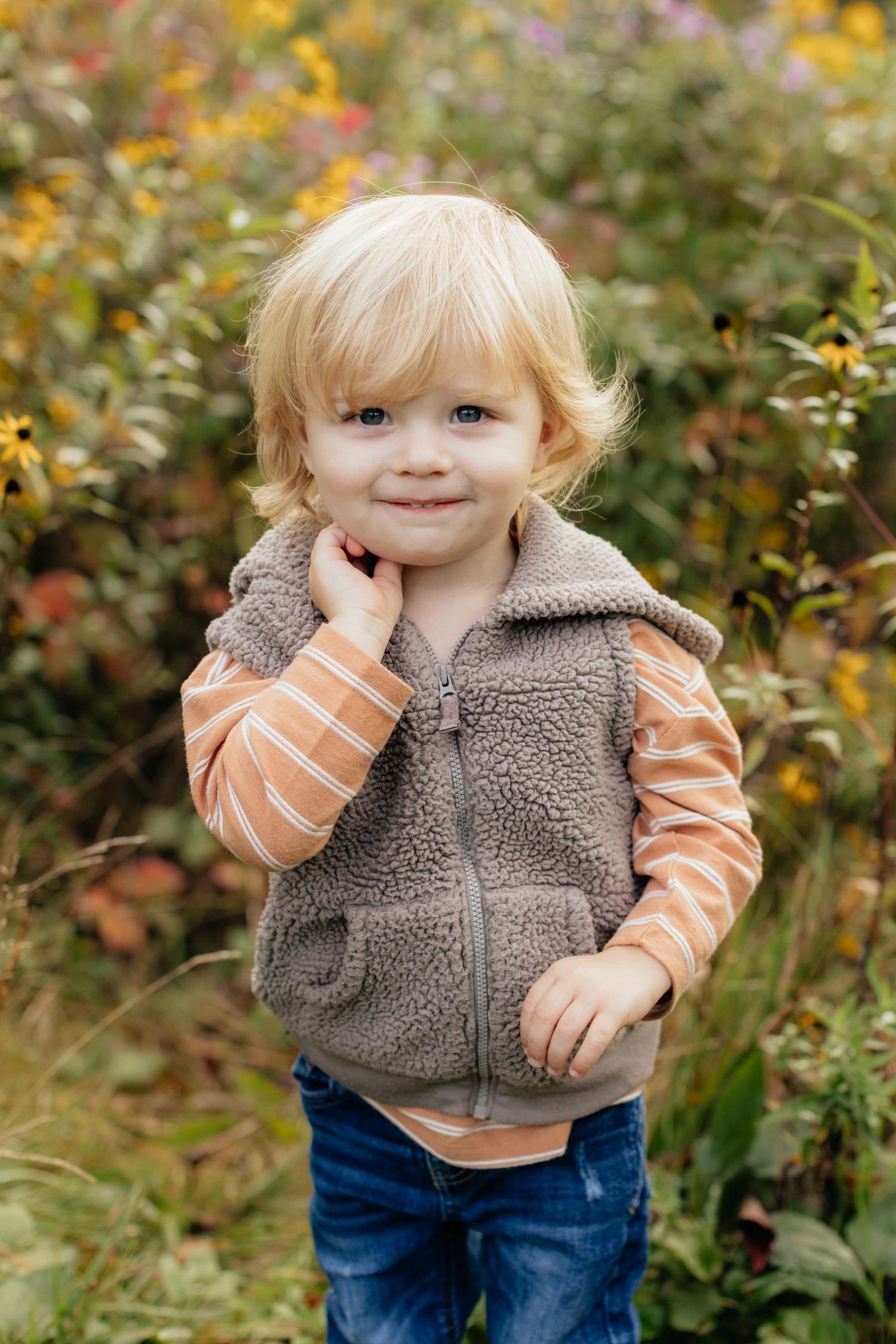 Toddler in striped shirt and fleece vest plays in a colorful autumn garden.