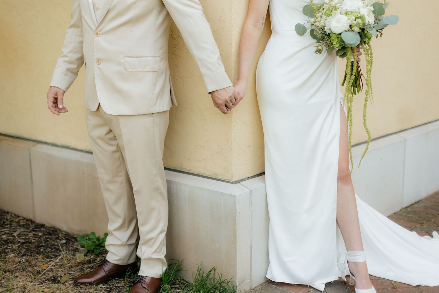 Bride and groom in coordinating white and beige outfits standing against yellow building wall.