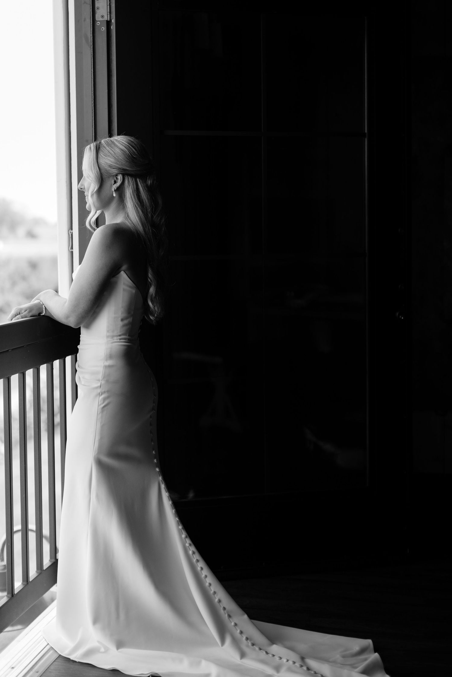Elegant bride in a white wedding dress poses by a window in dramatic lighting.