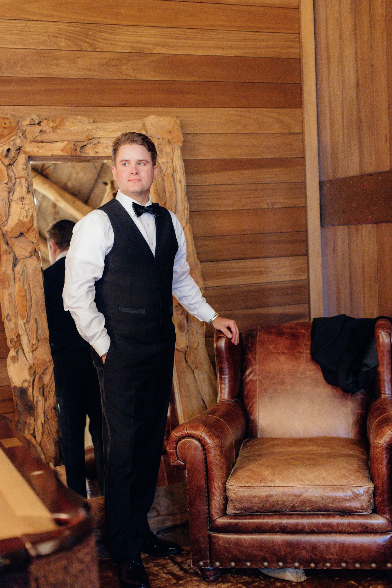A formal portrait of a groom in a tuxedo standing next to a leather armchair in a wooden room.