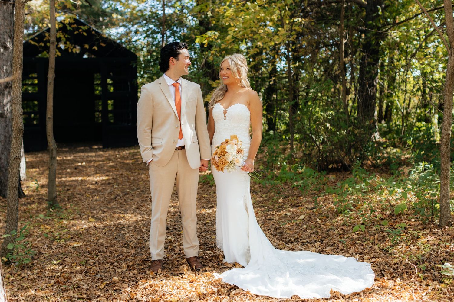 A wedding couple stands in dappled sunlight on fallen autumn leaves.