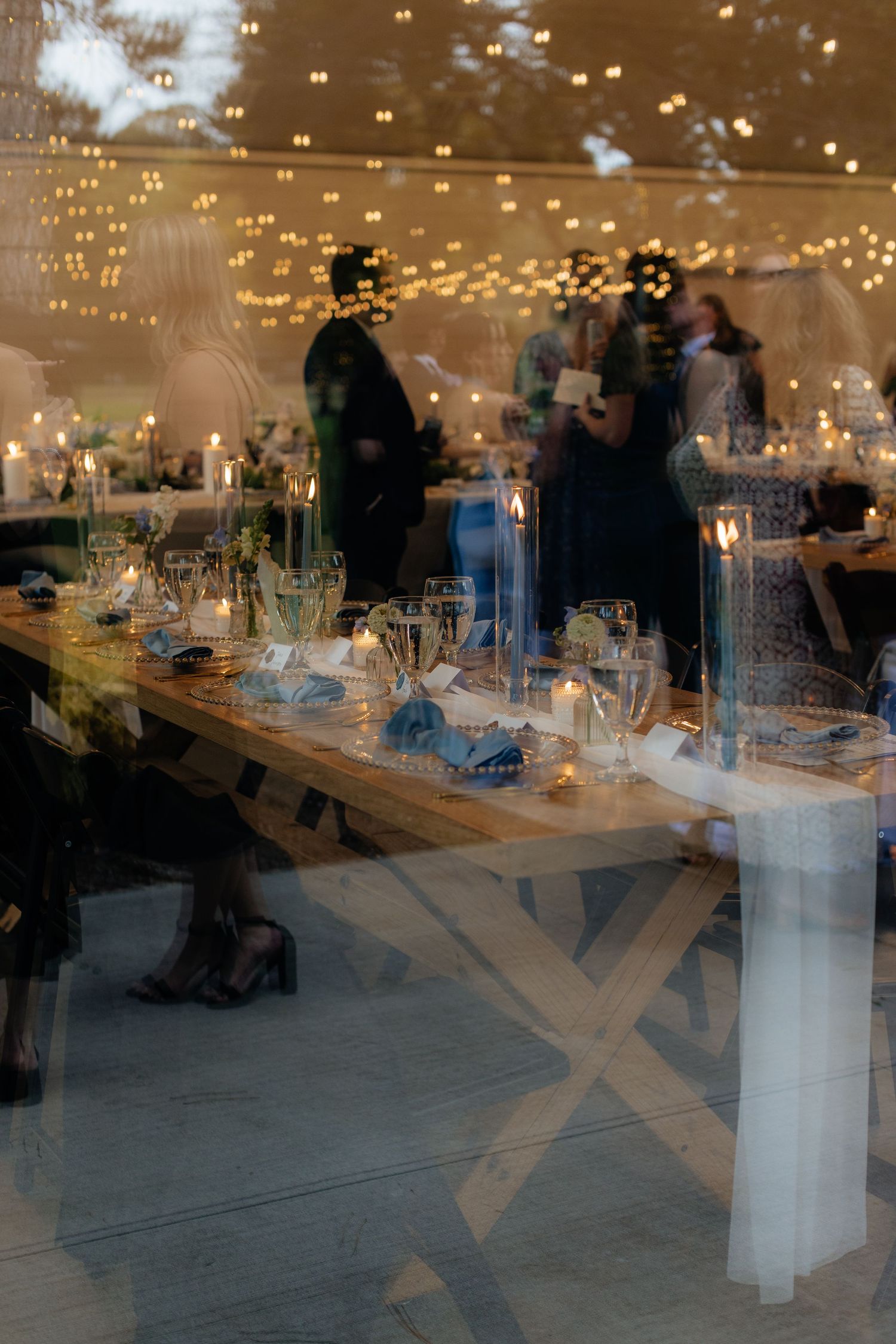 Wedding reception tables set with glowing candles and string lights reflecting in window.