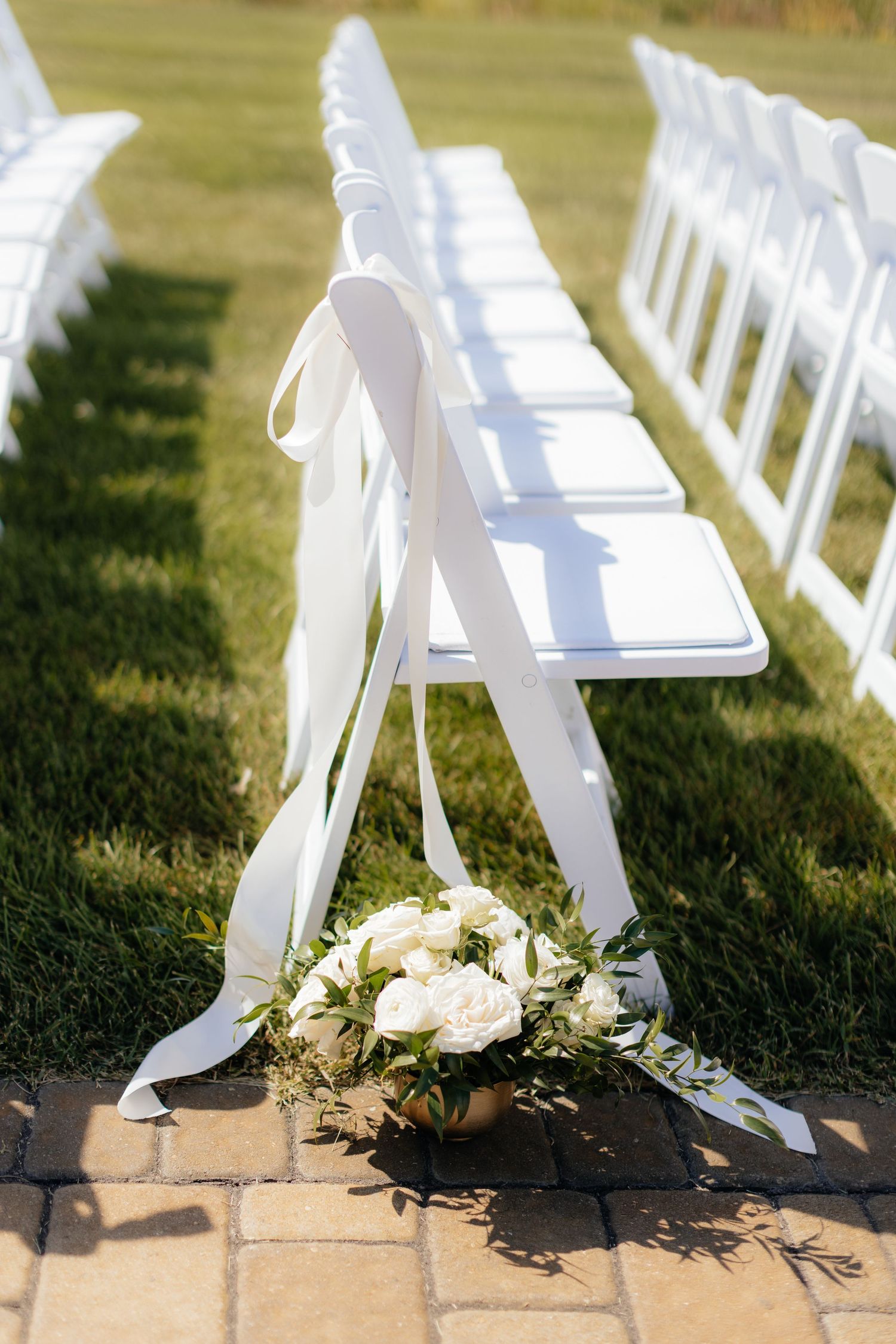 White folding chairs lined up for outdoor wedding ceremony with floral arrangement on aisle.