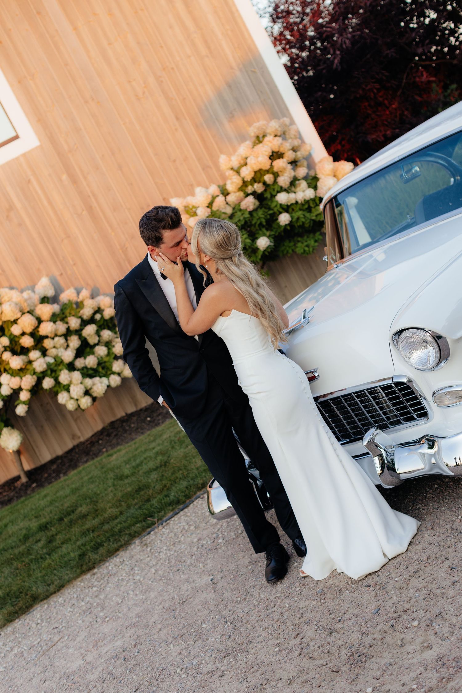 Romantic wedding couple embraces beside vintage white car with floral decorations.