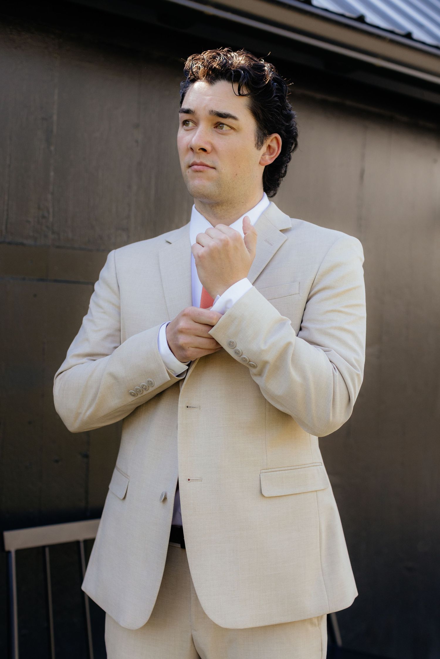 A person in a cream colored suit adjusts their tie while standing against a dark wall.