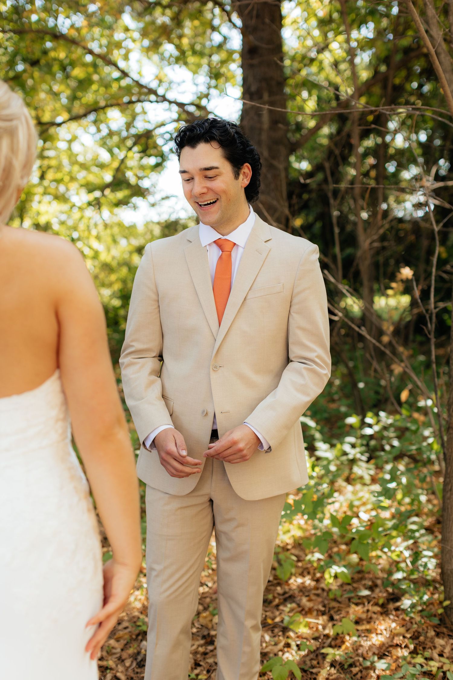First look wedding moment in a wooded setting with groom in beige suit and orange tie.