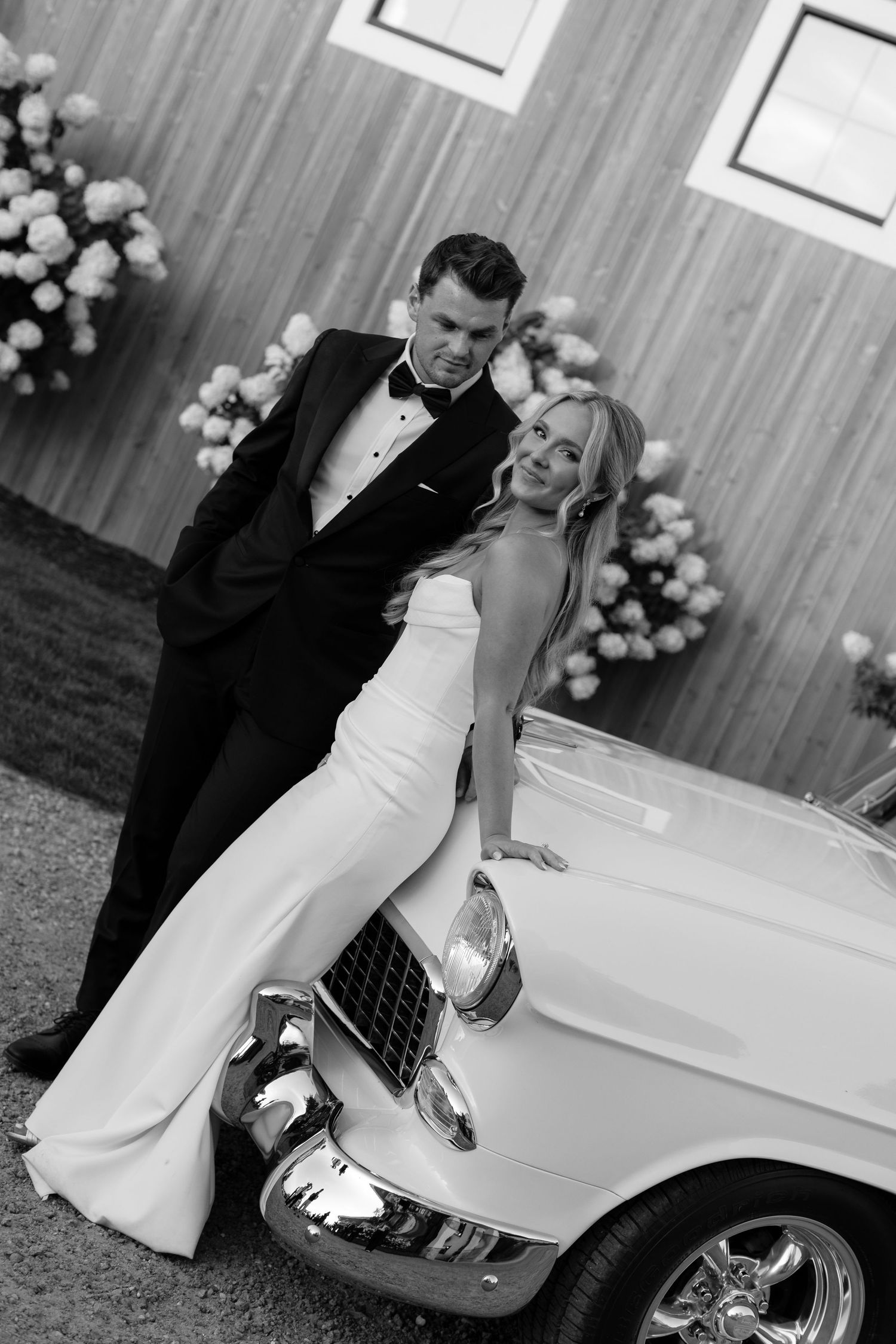Black and white portrait of couple posing against vintage Chevrolet at their wedding.