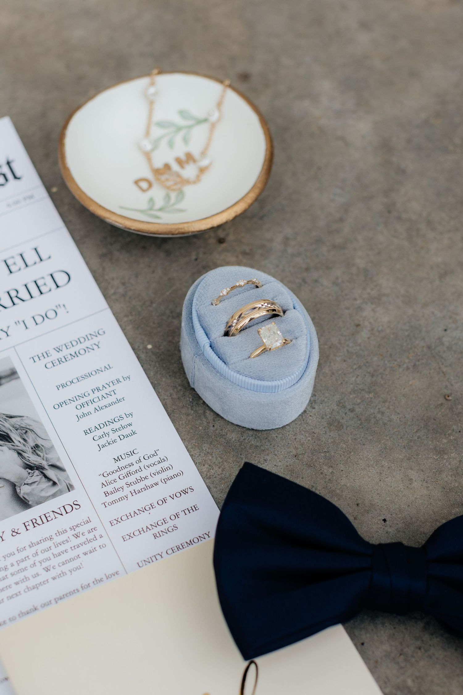 Wedding rings displayed in a light blue velvet box next to a navy bowtie and decorative dish.