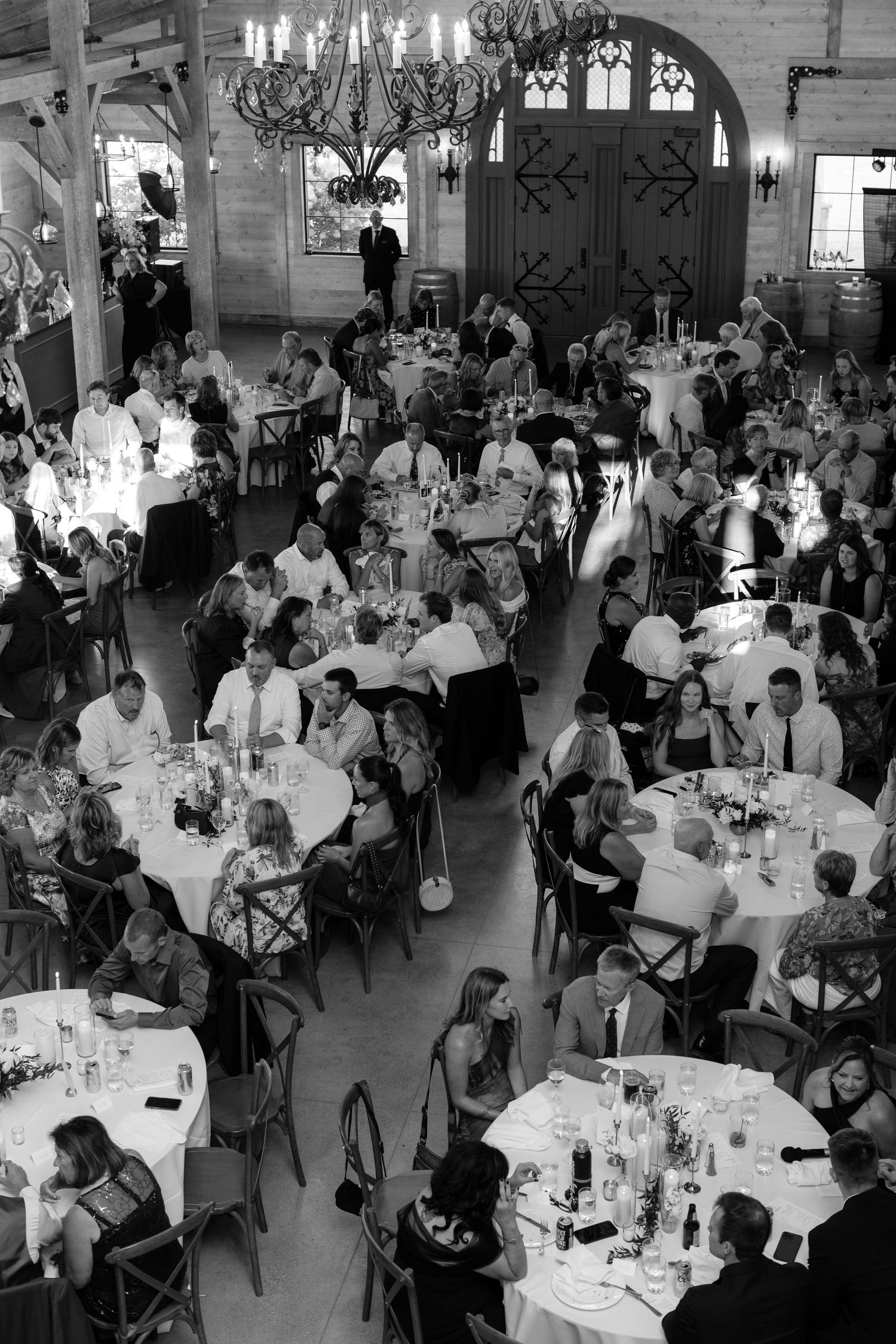 An elegant wedding reception hall filled with guests dining at round tables under crystal chandeliers.