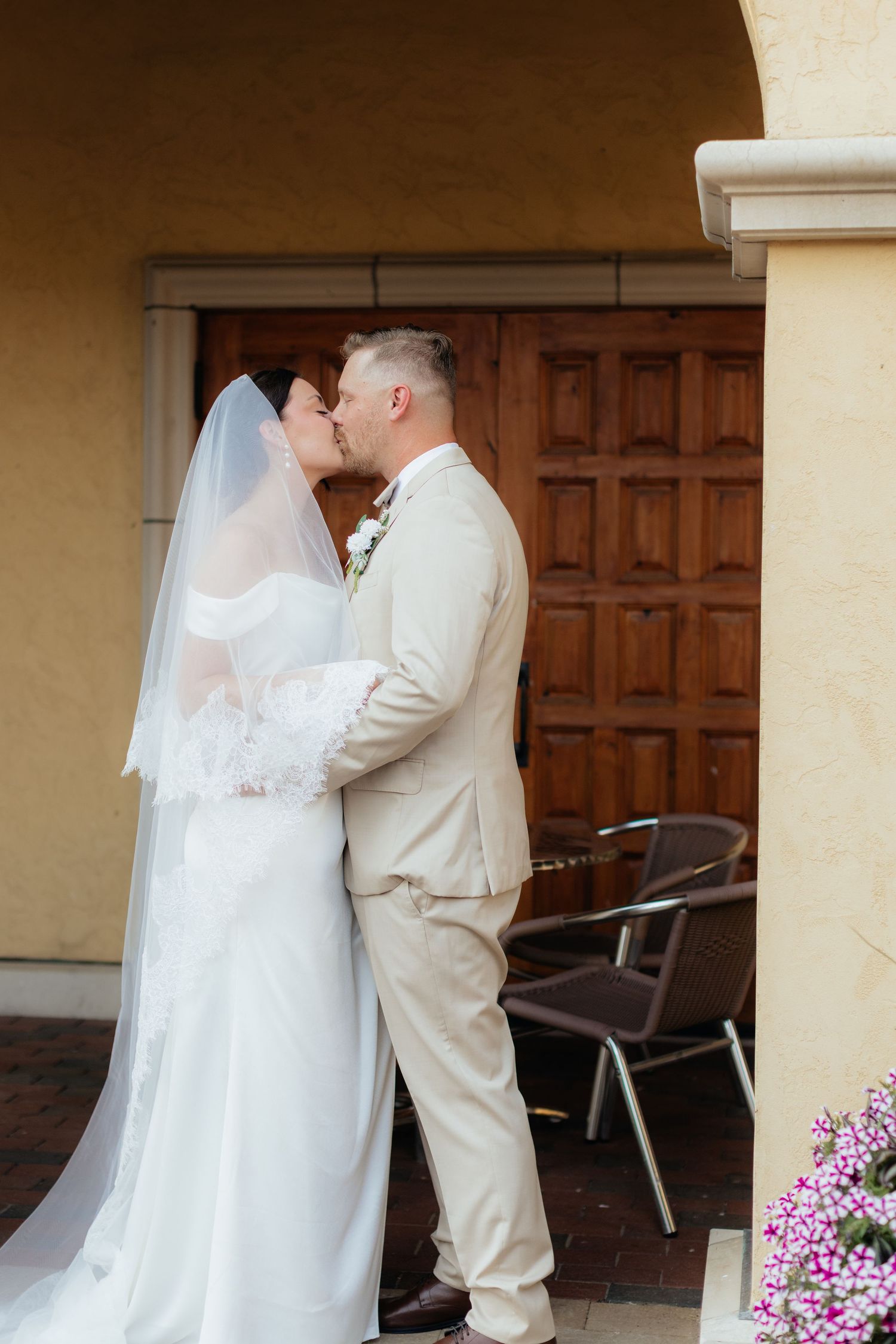 Wedding couple shares intimate moment under covered walkway with wooden doors and pink flowers.