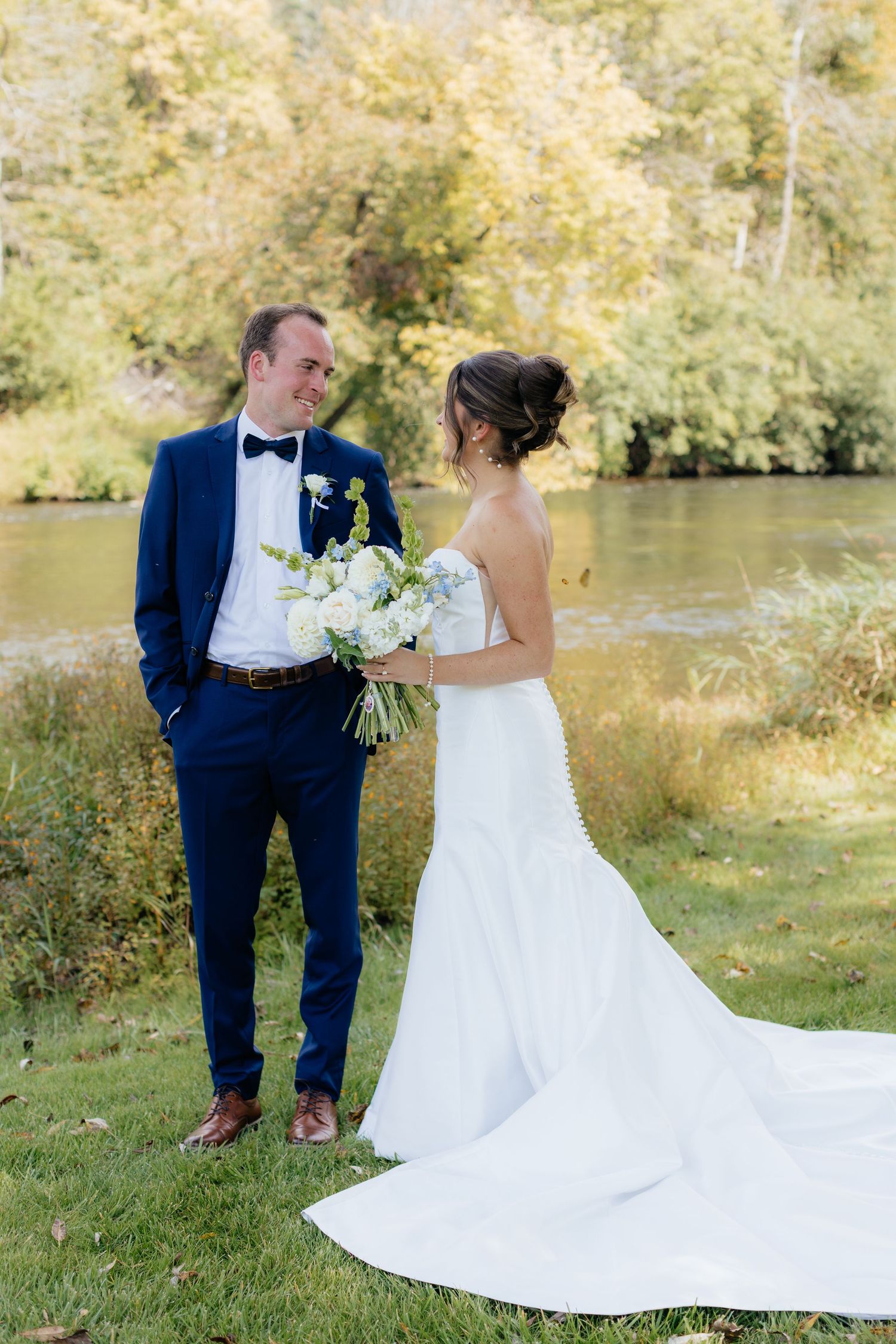 Wedding couple poses by riverside with bride's flowing white dress trailing on grass.