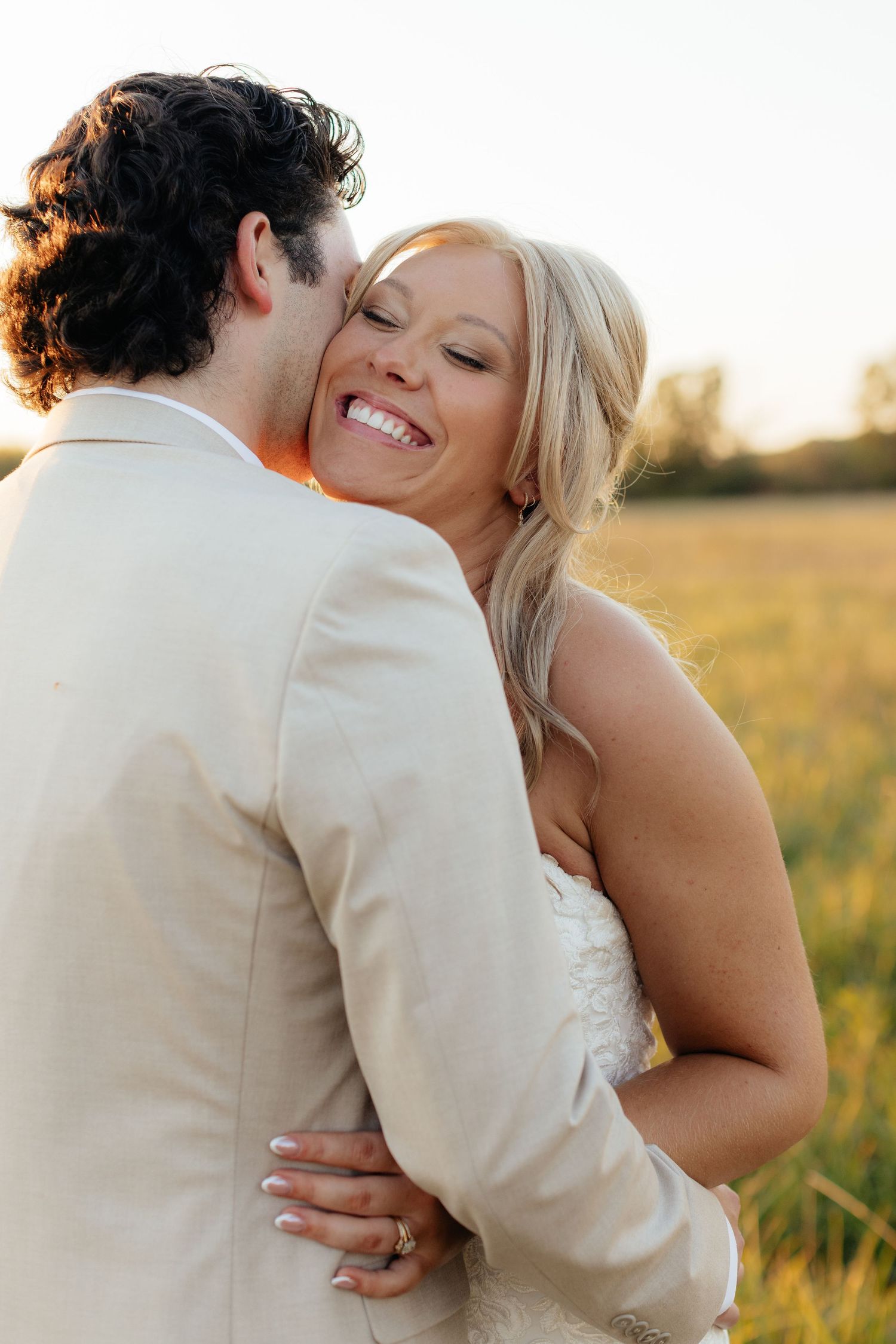 A romantic wedding couple embraces in a golden field at sunset.