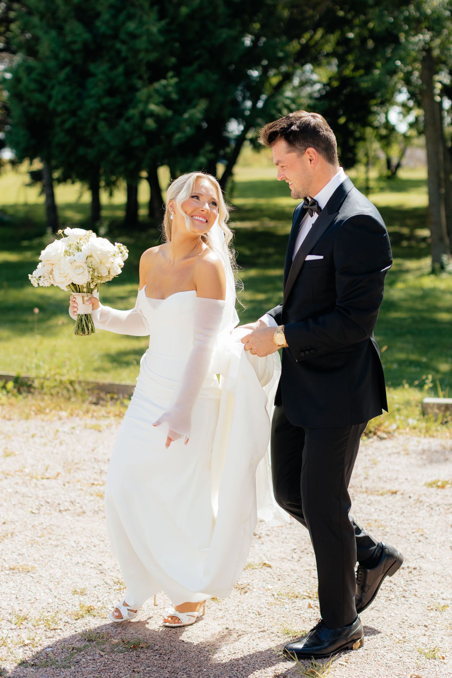 Playful wedding couple dances on gravel path surrounded by green trees.