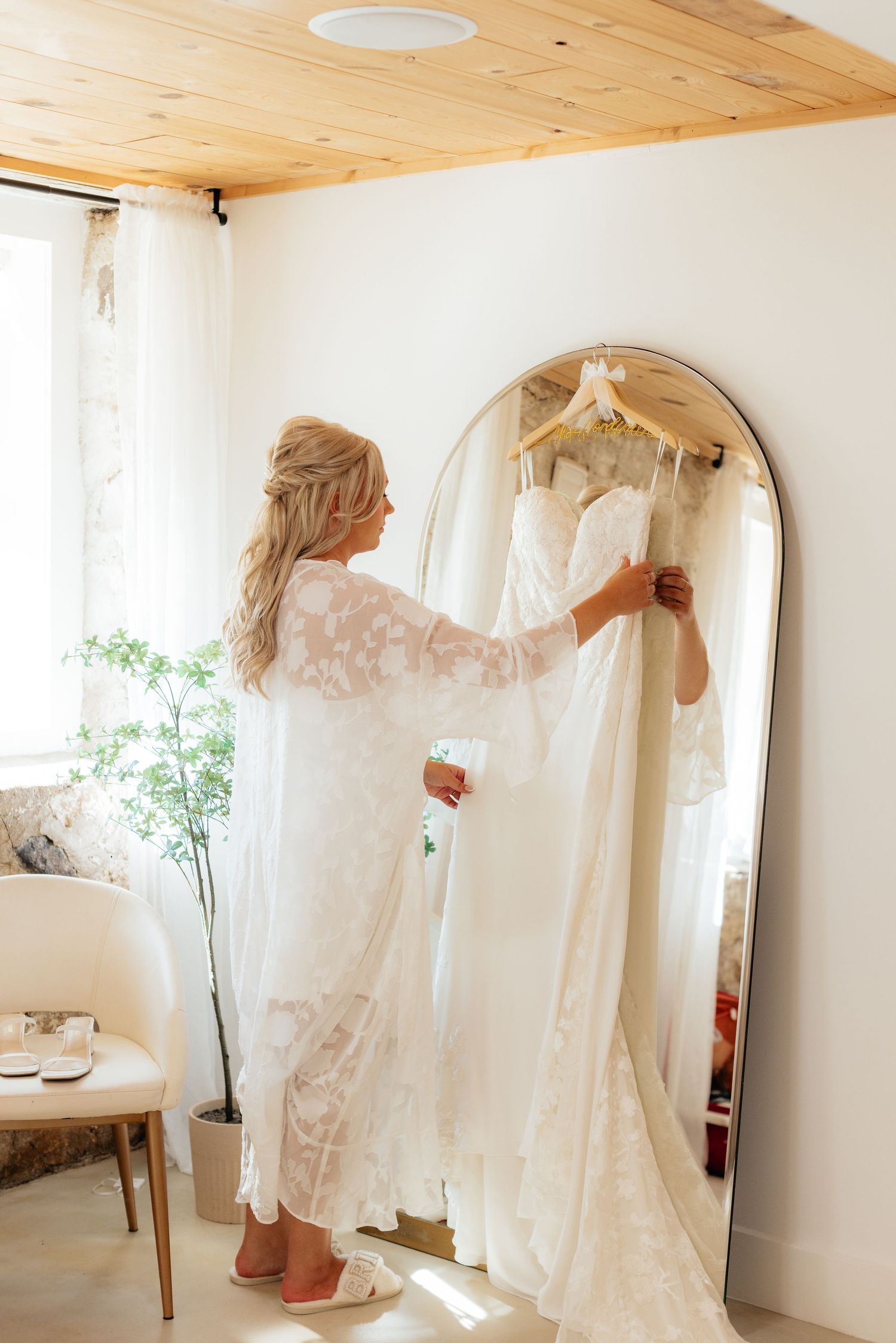 A wedding dress is being adjusted in a minimalist white room with wooden ceiling.