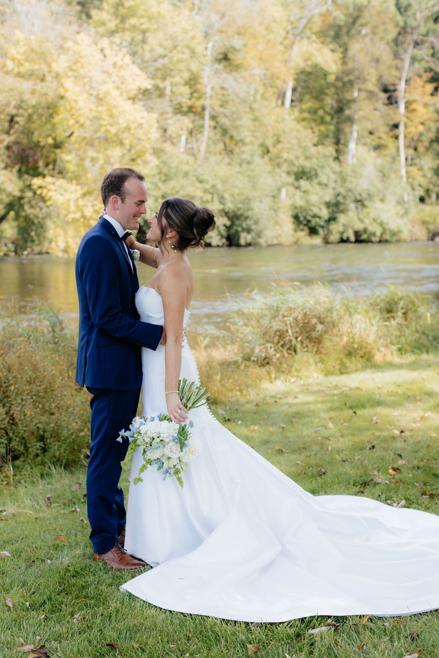 Romantic wedding portraits by a river with autumn trees in the background.