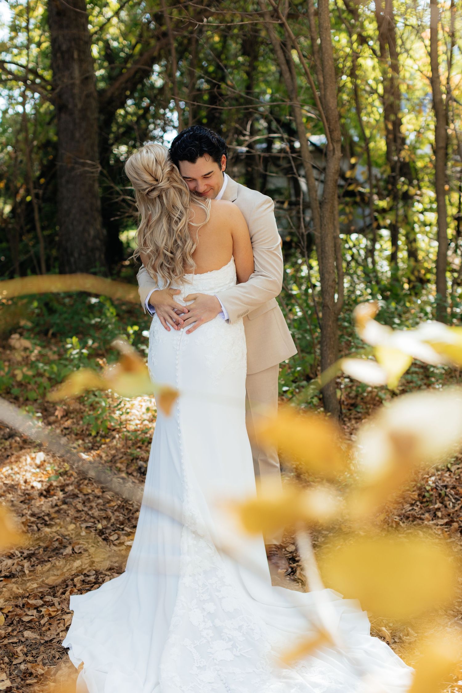 Romantic wedding portrait in sunlit forest with flowing white dress and beige suit.