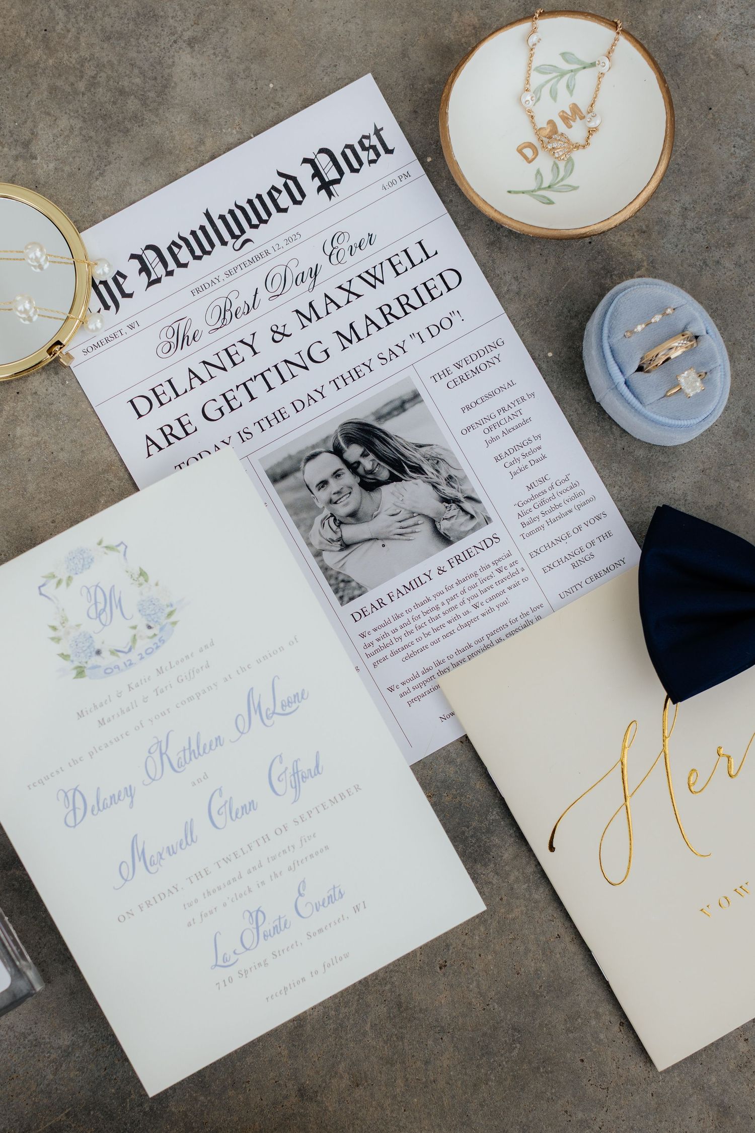 Wedding details including custom newspaper announcement, vow books, and rings on concrete surface.