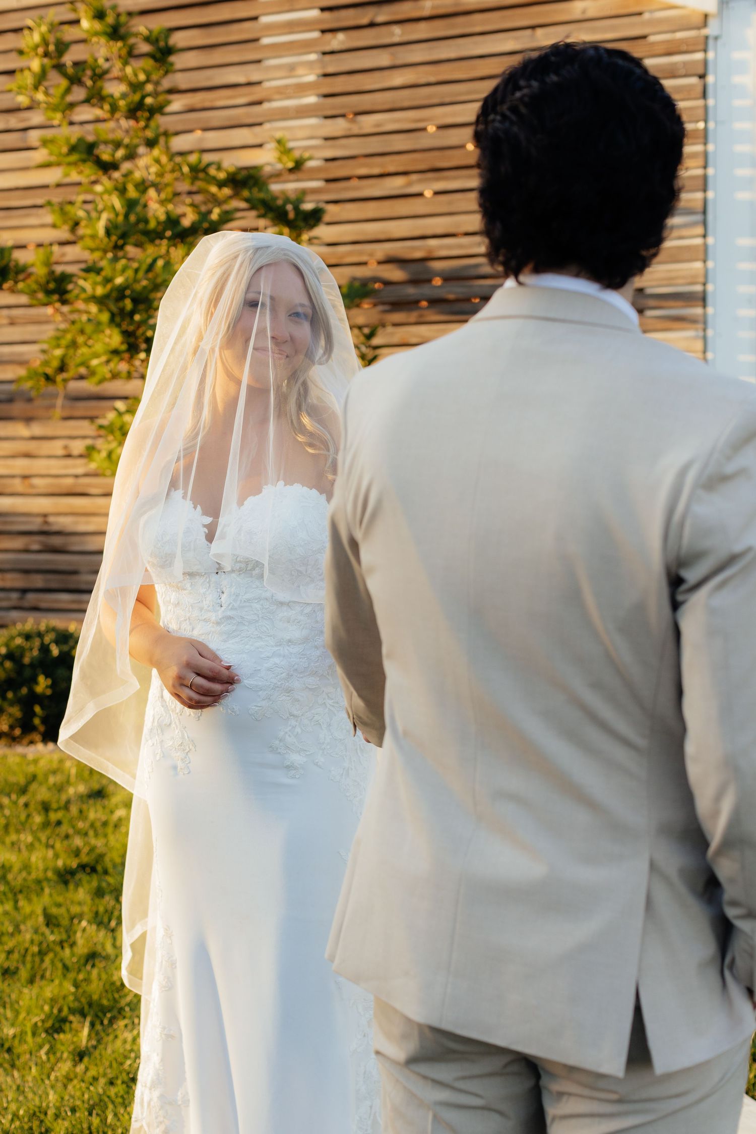Couple in wedding attire walking together through a sunlit outdoor setting with wooden paneled wall.
