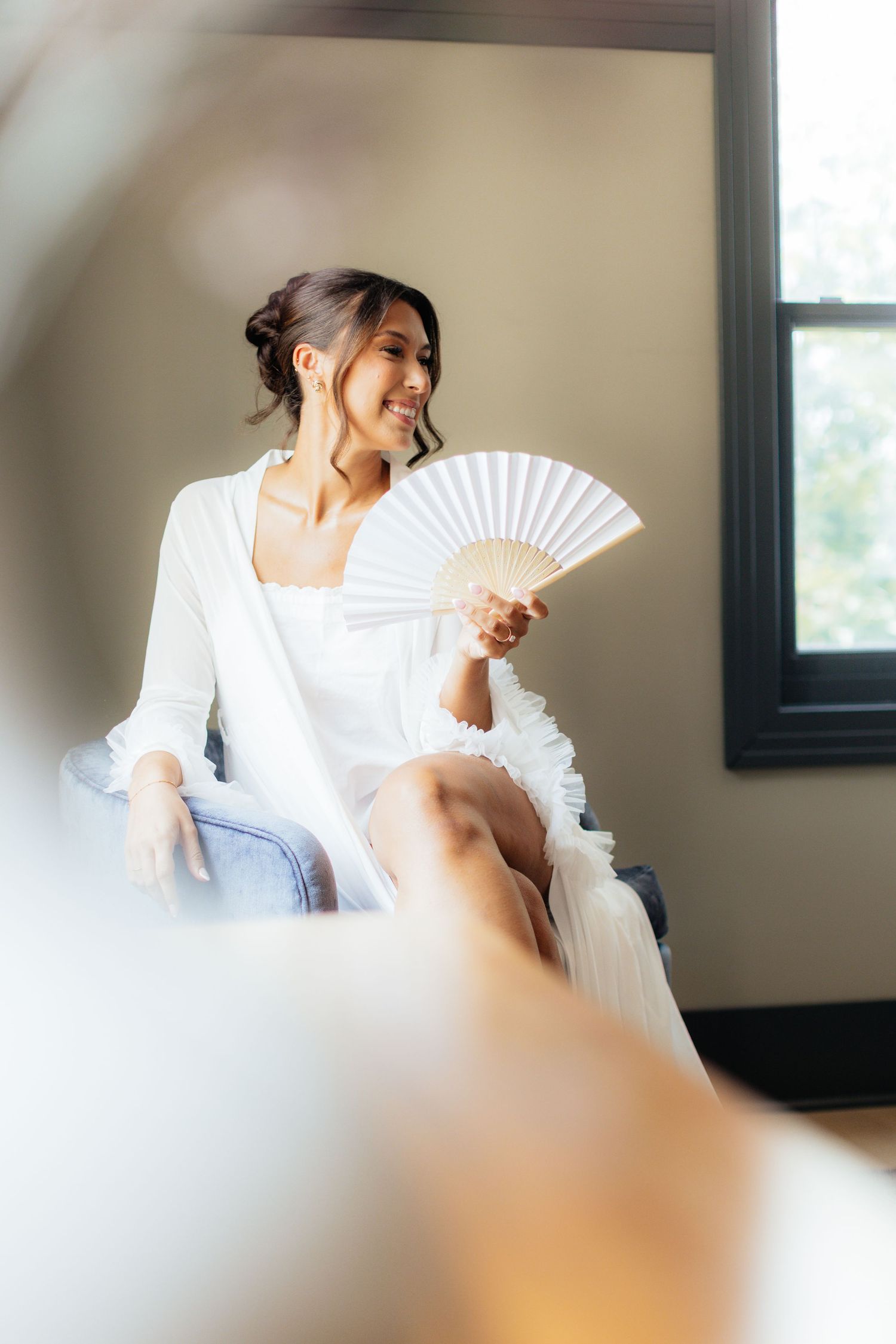 Woman in white dress poses elegantly with a decorative fan in a minimalist indoor setting.