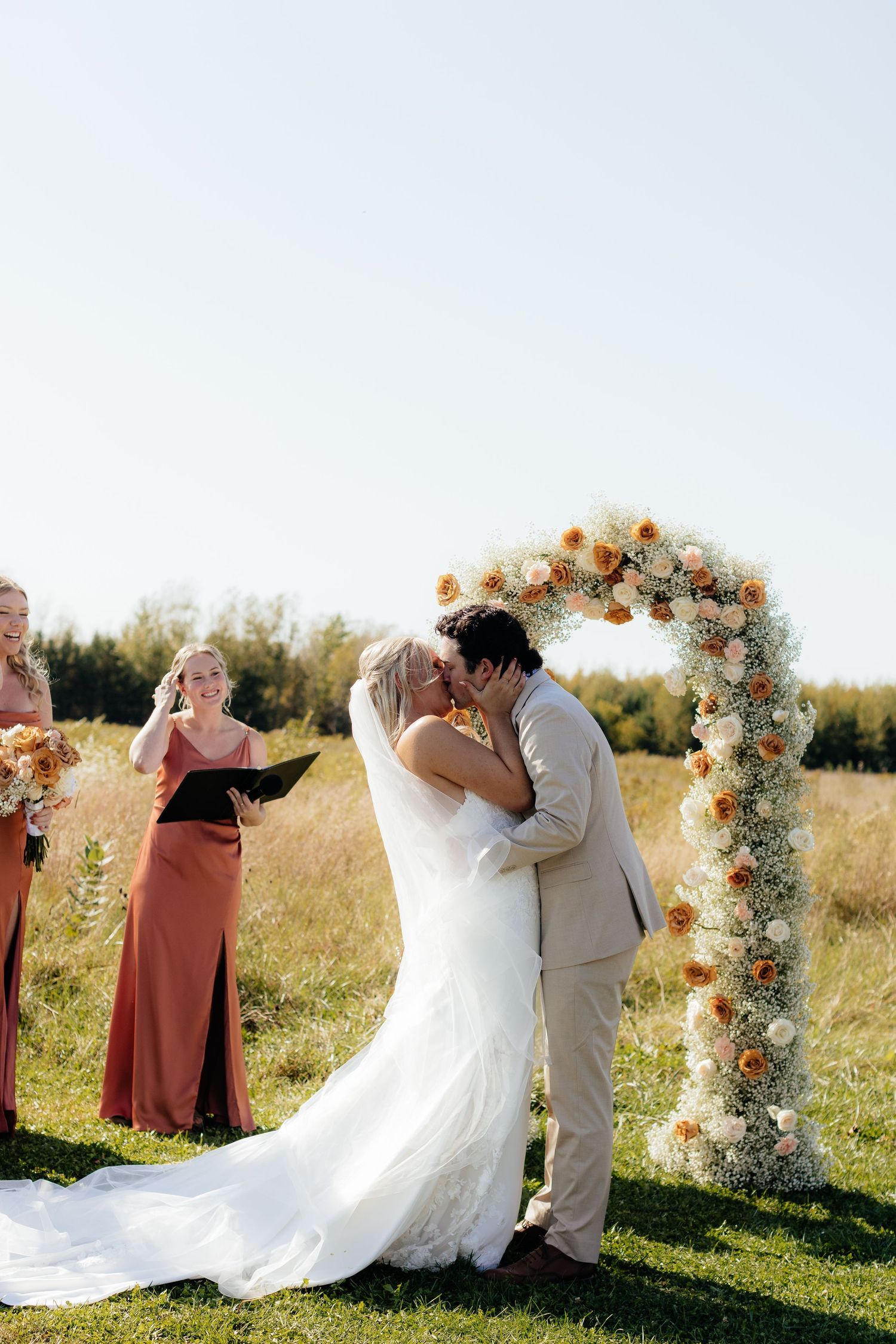 Wedding ceremony under a floral arch in a rustic outdoor setting during sunset.