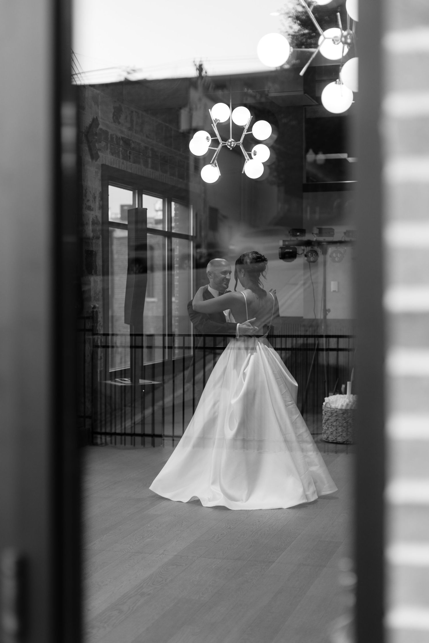A black and white reflection shows dancers through window panes with hanging lights above.