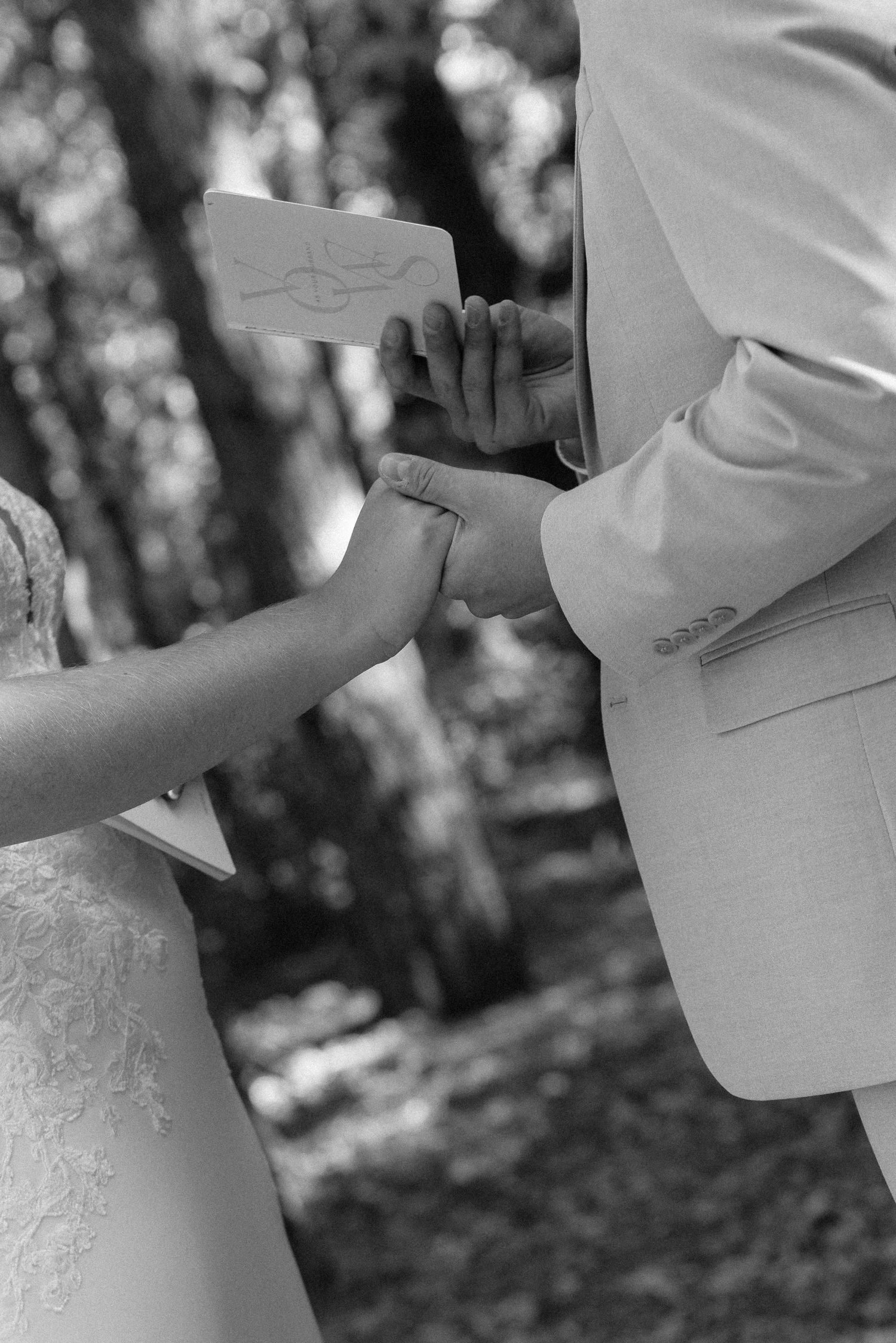 Black and white close-up of hands exchanging wedding vows in outdoor ceremony.