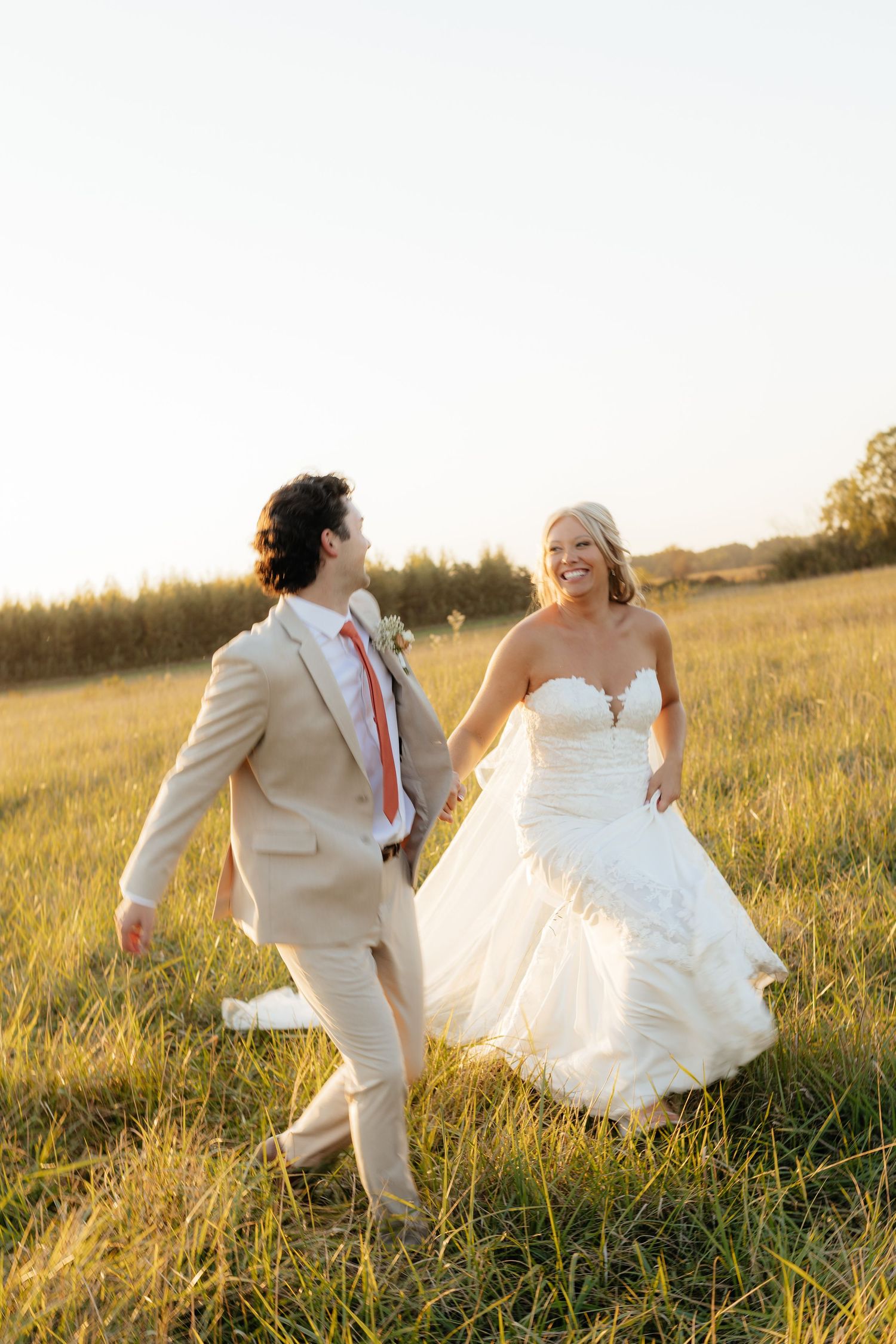 Playful newlyweds running through tall grass field at sunset.