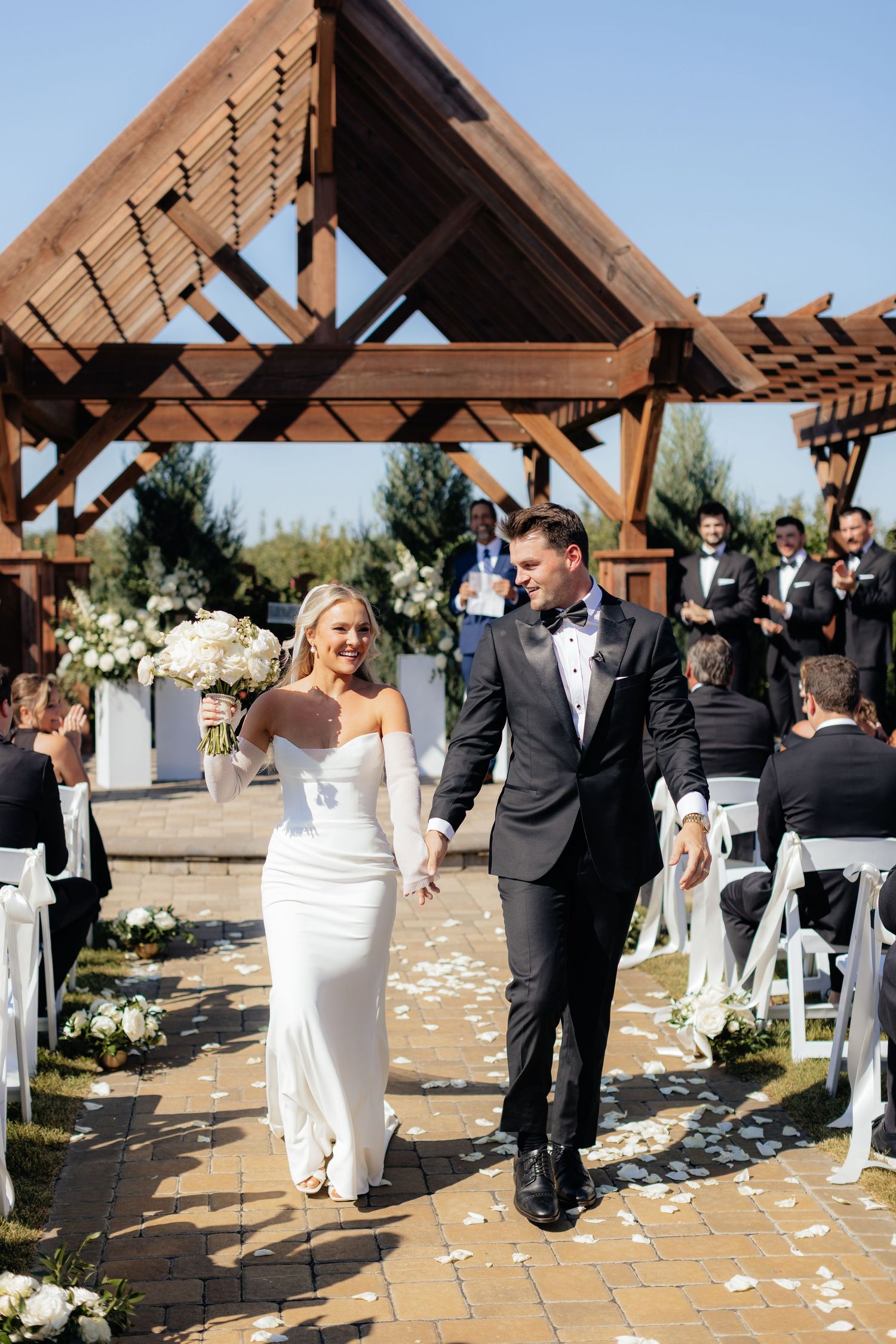 Wedding ceremony recessional under wooden pergola with white chairs and scattered petals.