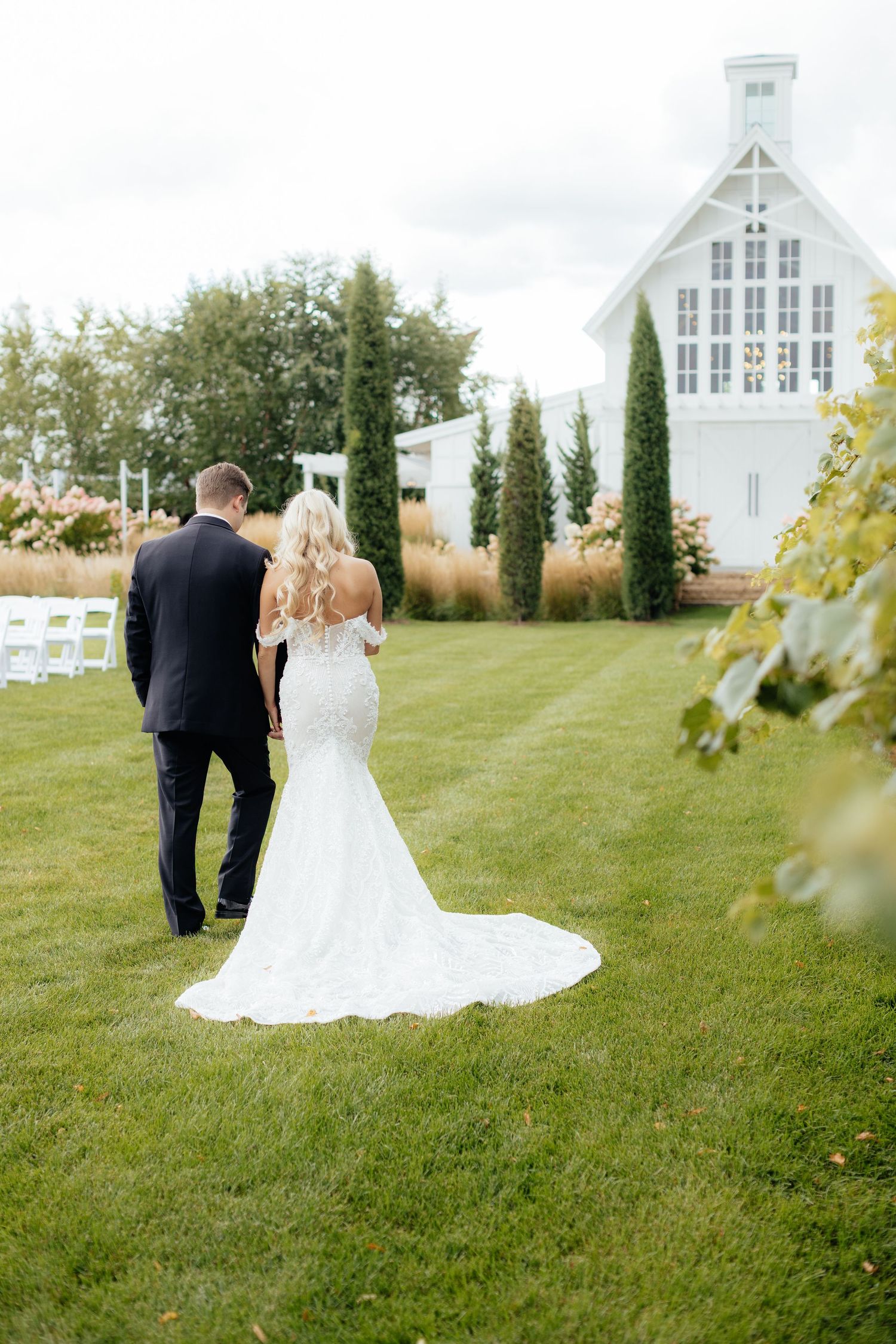 Wedding couple poses on grassy lawn with white chapel and vineyard in background.